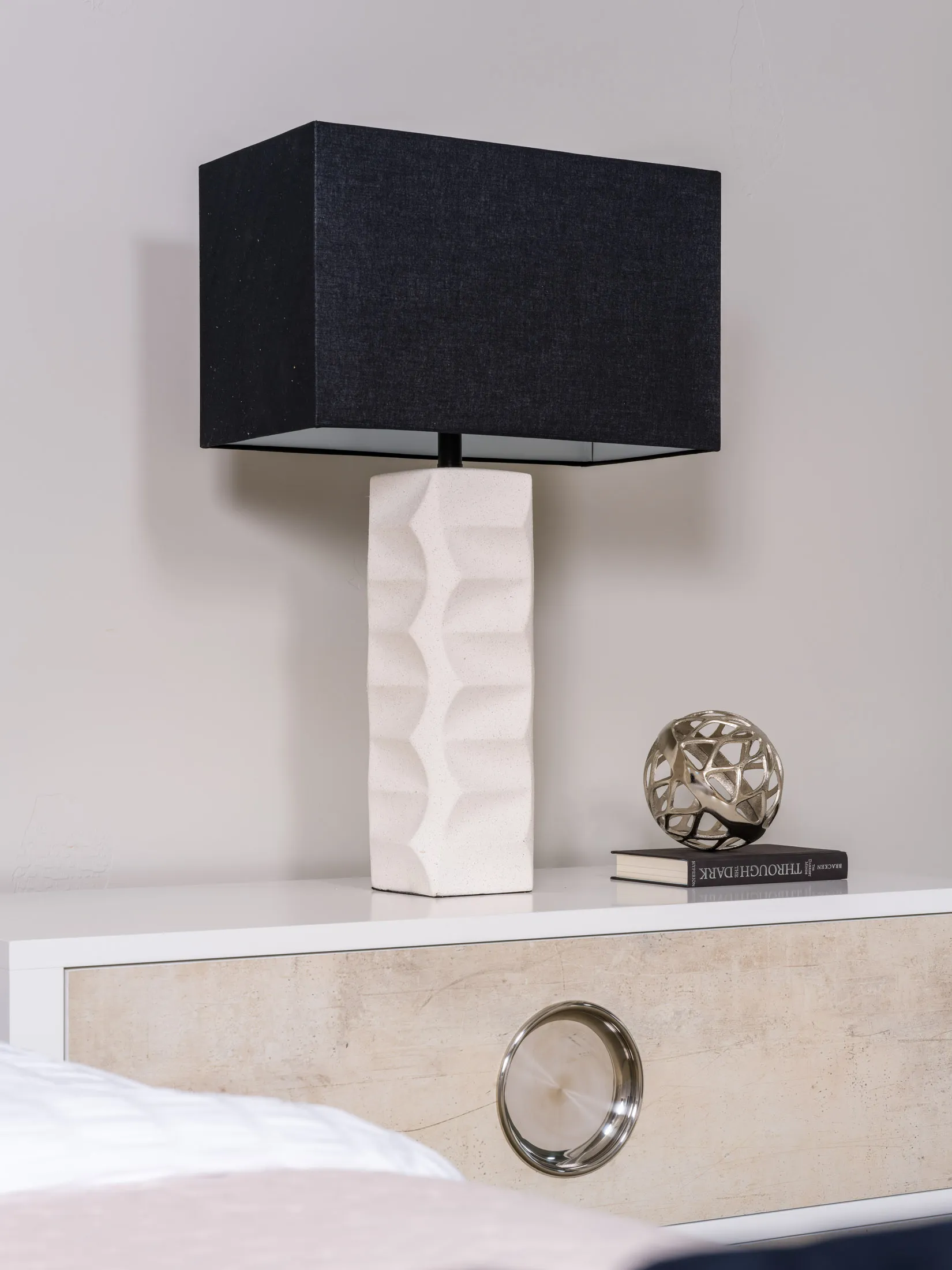 A textured white lamp with a black rectangular shade on a modern bedside table, accompanied by a silver decorative sphere and a book.