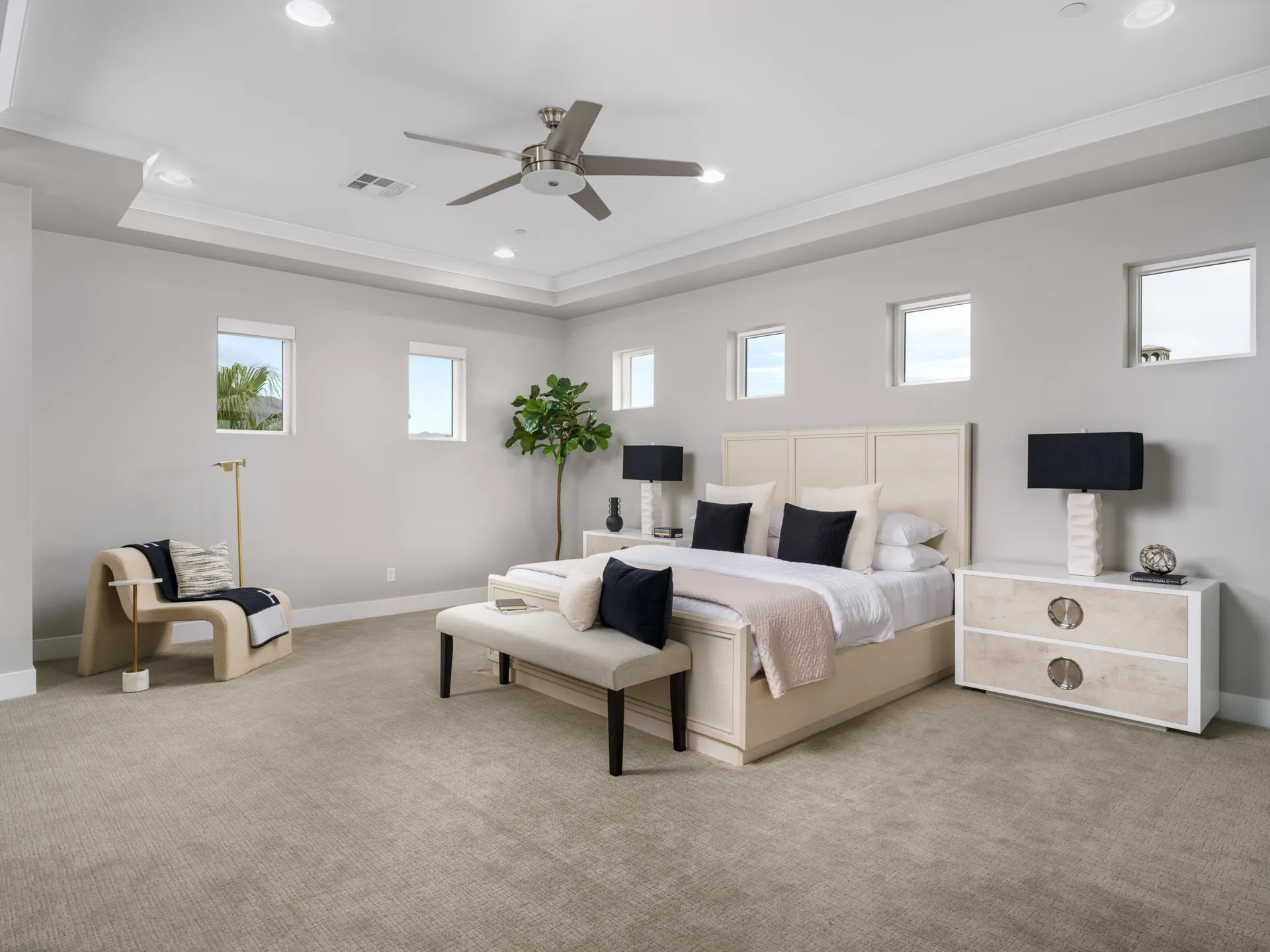 A large bedroom with a light wood bed, a neutral bench at the foot, black accent lamps on matching nightstands, and a modern chair in the corner.