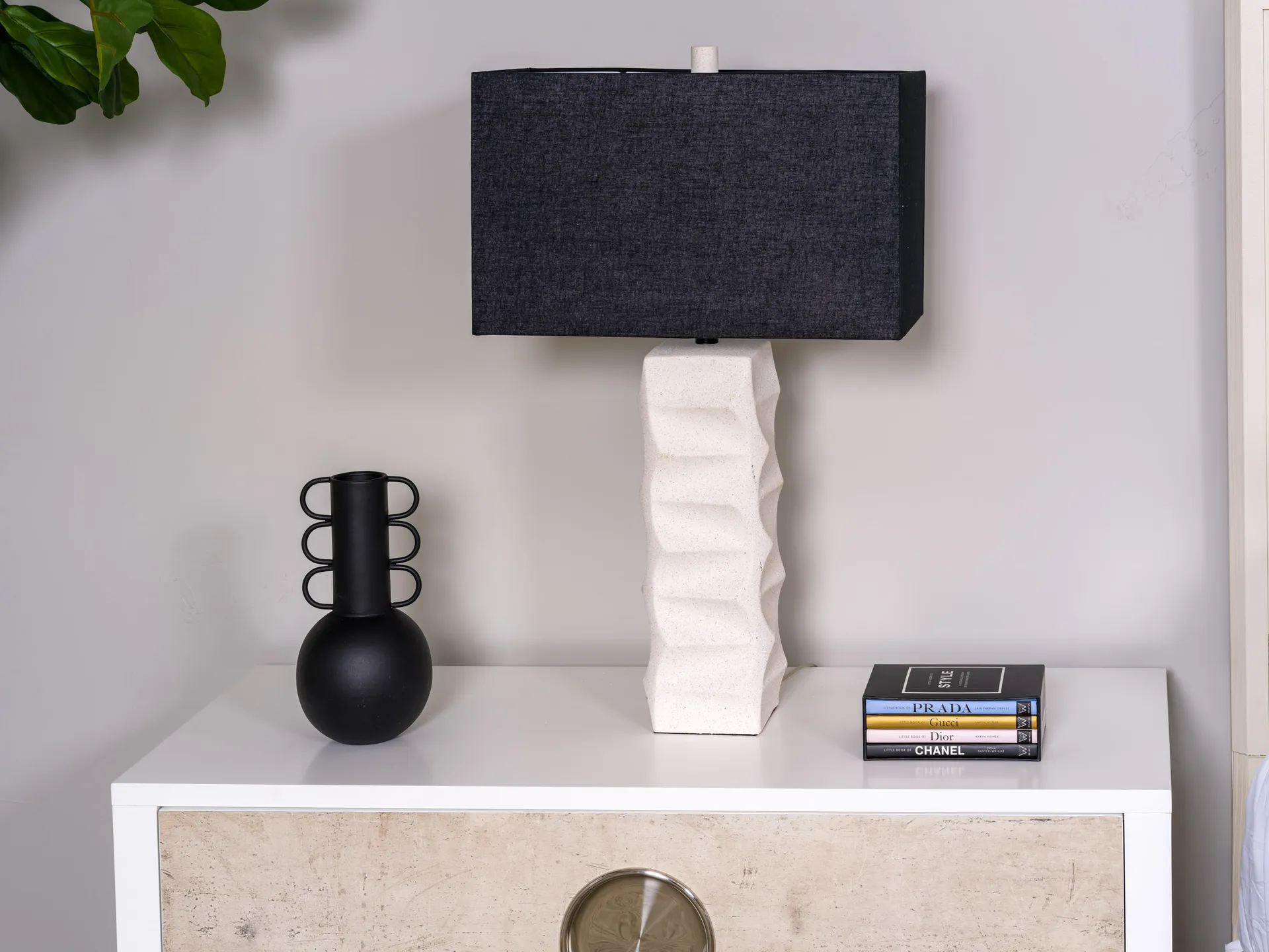 A white nightstand with a textured white lamp featuring a black rectangular shade, alongside a black ring-handled vase and a small stack of fashion books.