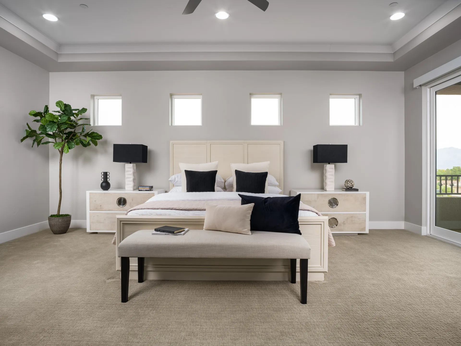 A spacious neutral bedroom with a light wood bed, black accent pillows, matching nightstands, and a fiddle leaf fig in one corner.