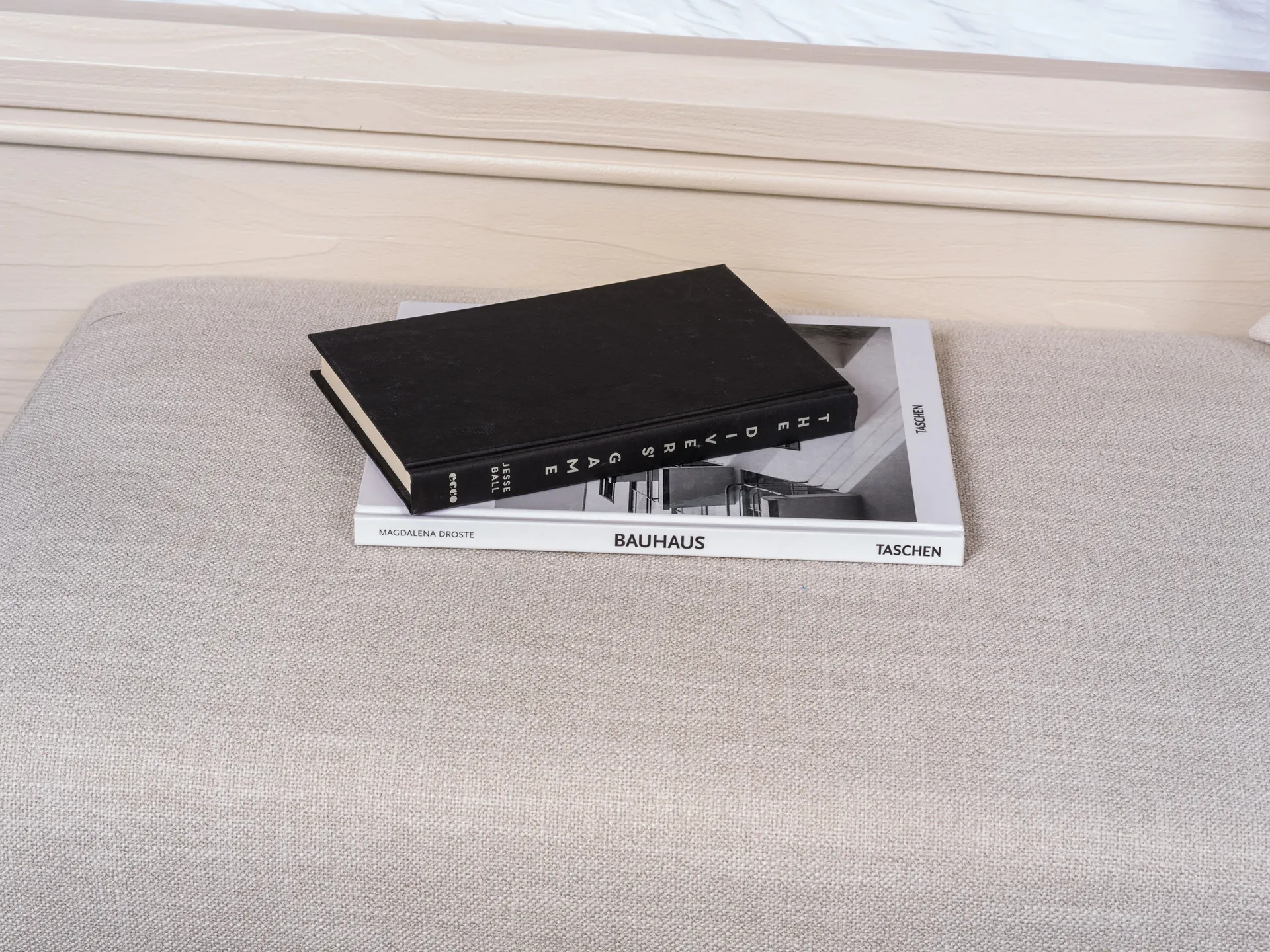 Two hardcover books, one black and one featuring Bauhaus imagery, placed on a neutral upholstered bench.