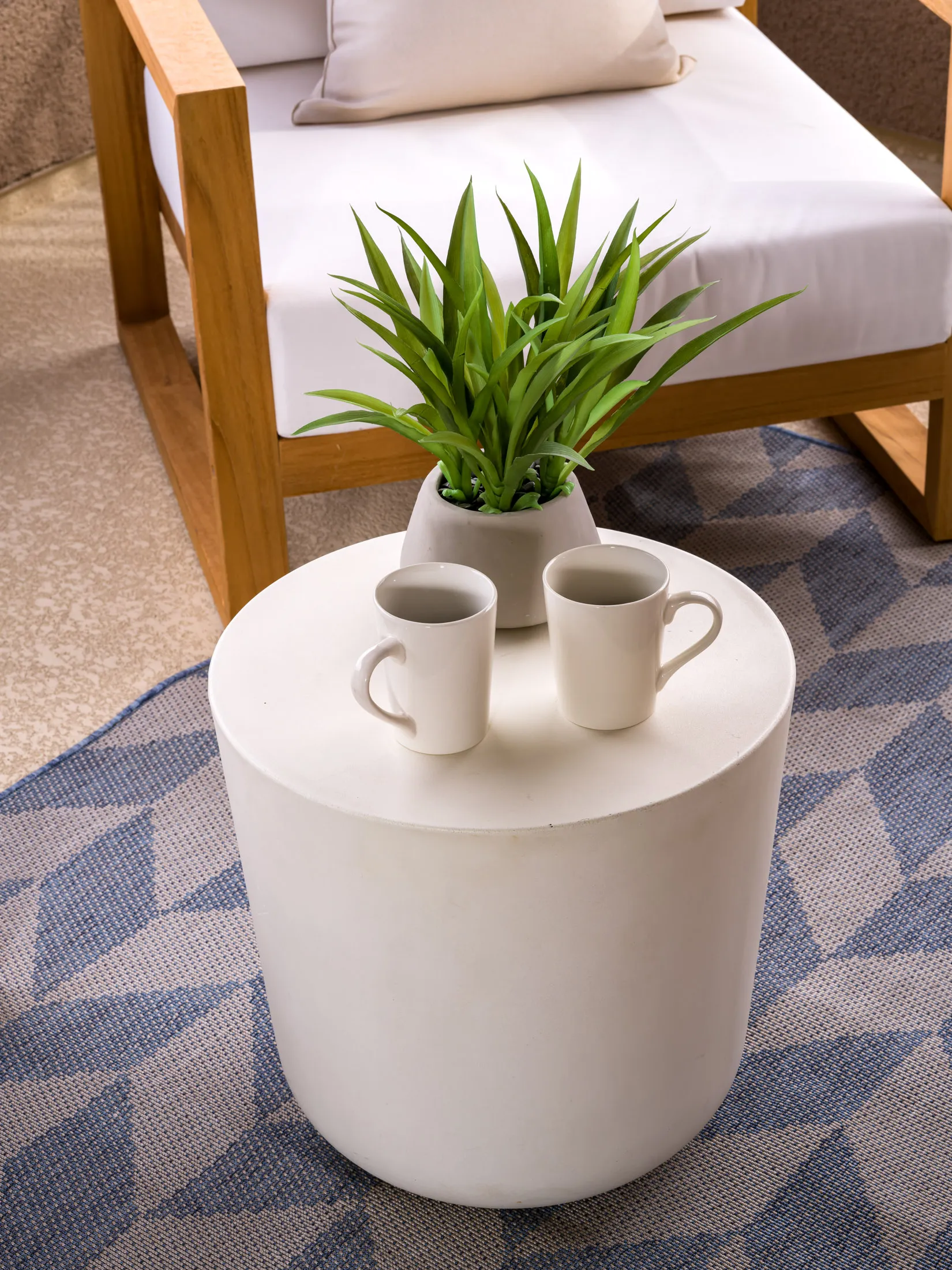 A white cylindrical side table with two coffee mugs and a small green plant, next to a wooden outdoor chair on a blue patterned rug.