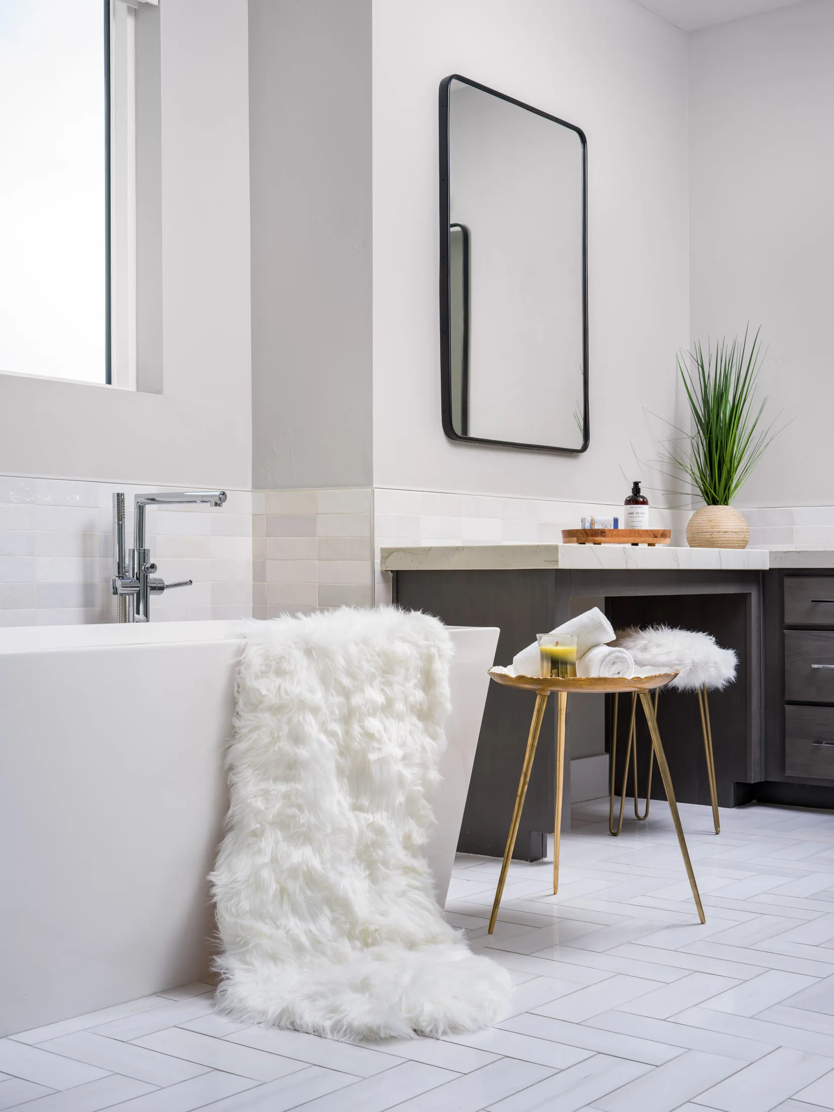 A sleek white bathtub with a faux-fur throw draped over the side, next to a gold accent table holding towels and a candle. A black-framed mirror and a small plant add modern touches.