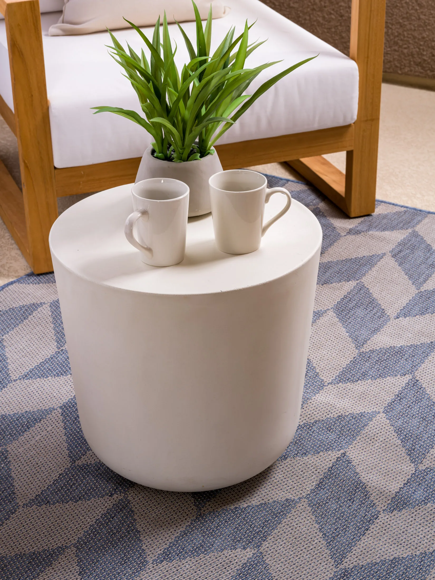 A cylindrical white table holding two coffee mugs and a small green plant, set next to a wooden outdoor chair on a geometric-patterned rug.
