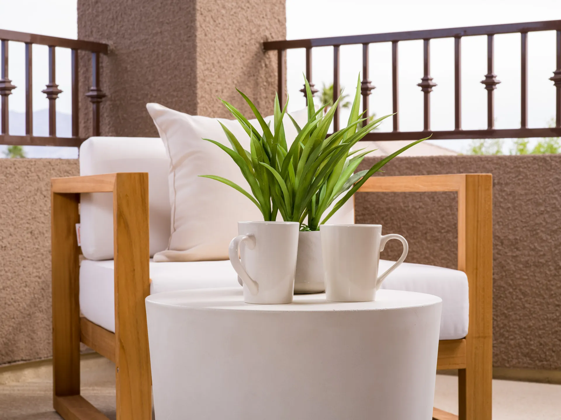 A small white table with two coffee mugs and a potted plant, set beside a wooden outdoor chair on a covered balcony.