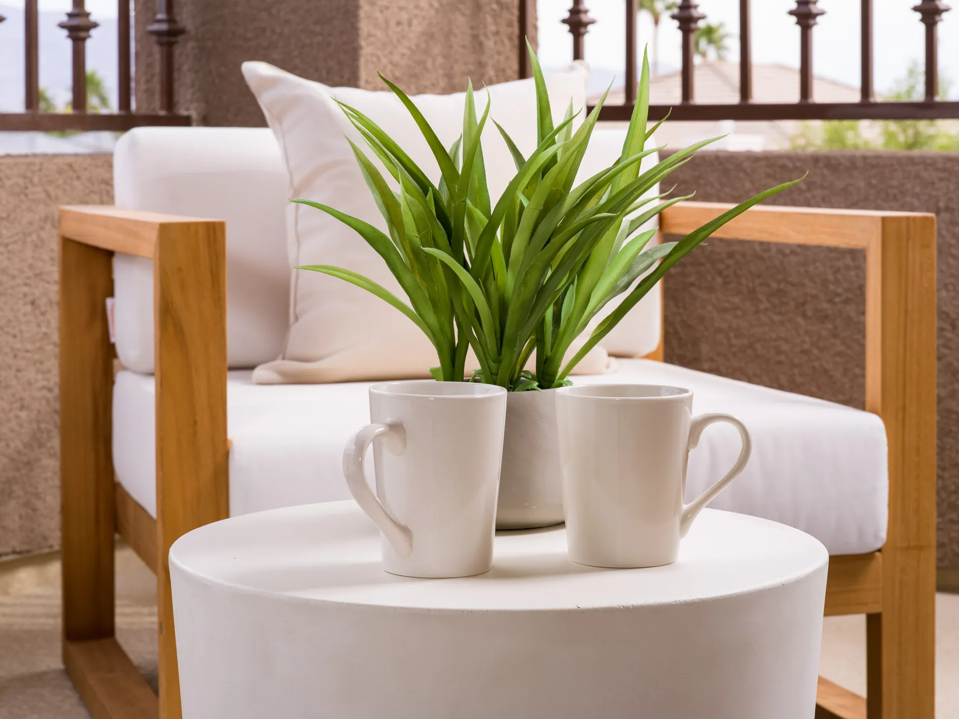 Two white mugs and a small green plant on a round white table, with a wooden outdoor chair in the background.