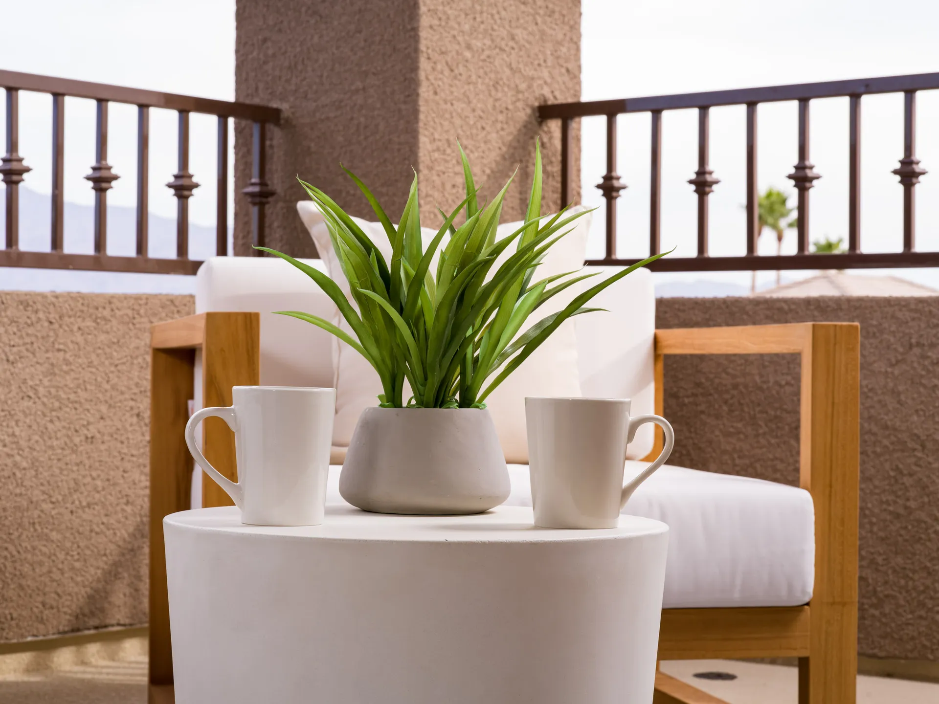 A small white table on a covered balcony, holding a potted plant and two white coffee mugs, with wooden outdoor seating in the background.