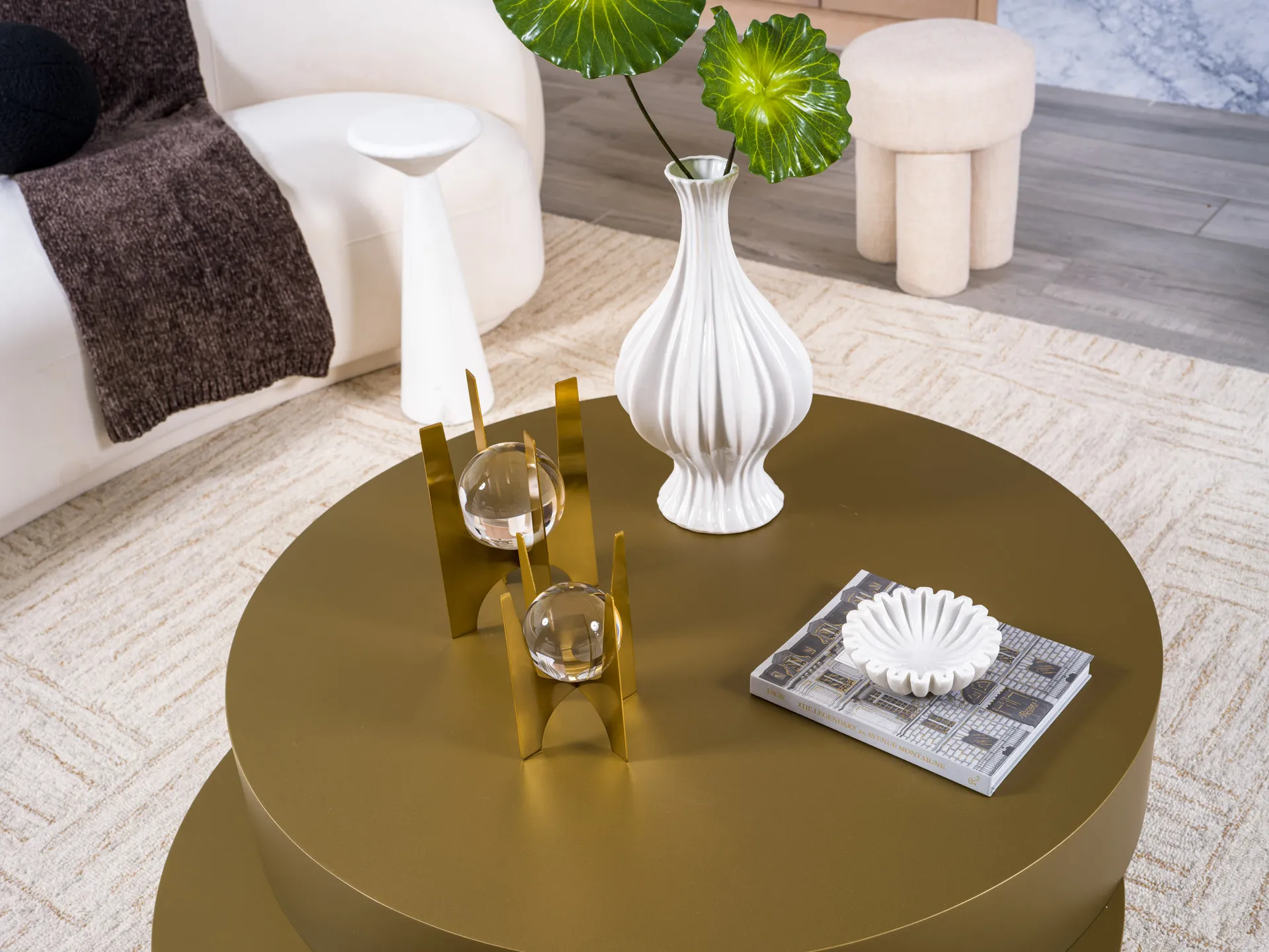 A round gold coffee table with a white fluted vase holding green leaves, a gold sculpture supporting glass orbs, and a small white dish on a hardcover book.