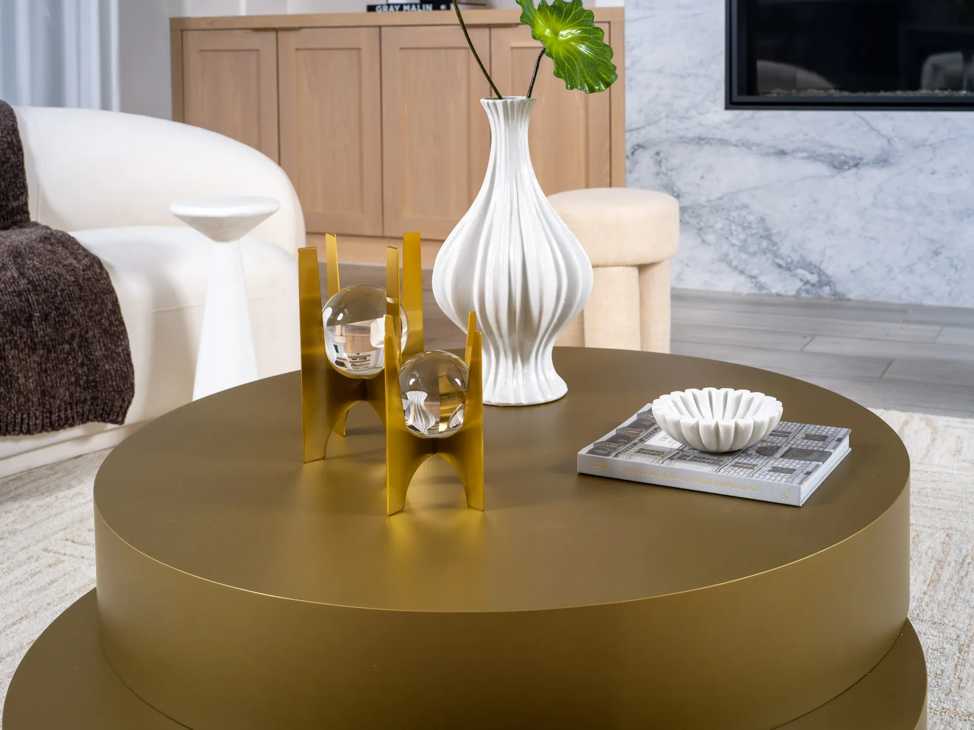 A round gold coffee table with a white fluted vase, a gold sculpture holding glass orbs, and a small white dish on a hardcover book.