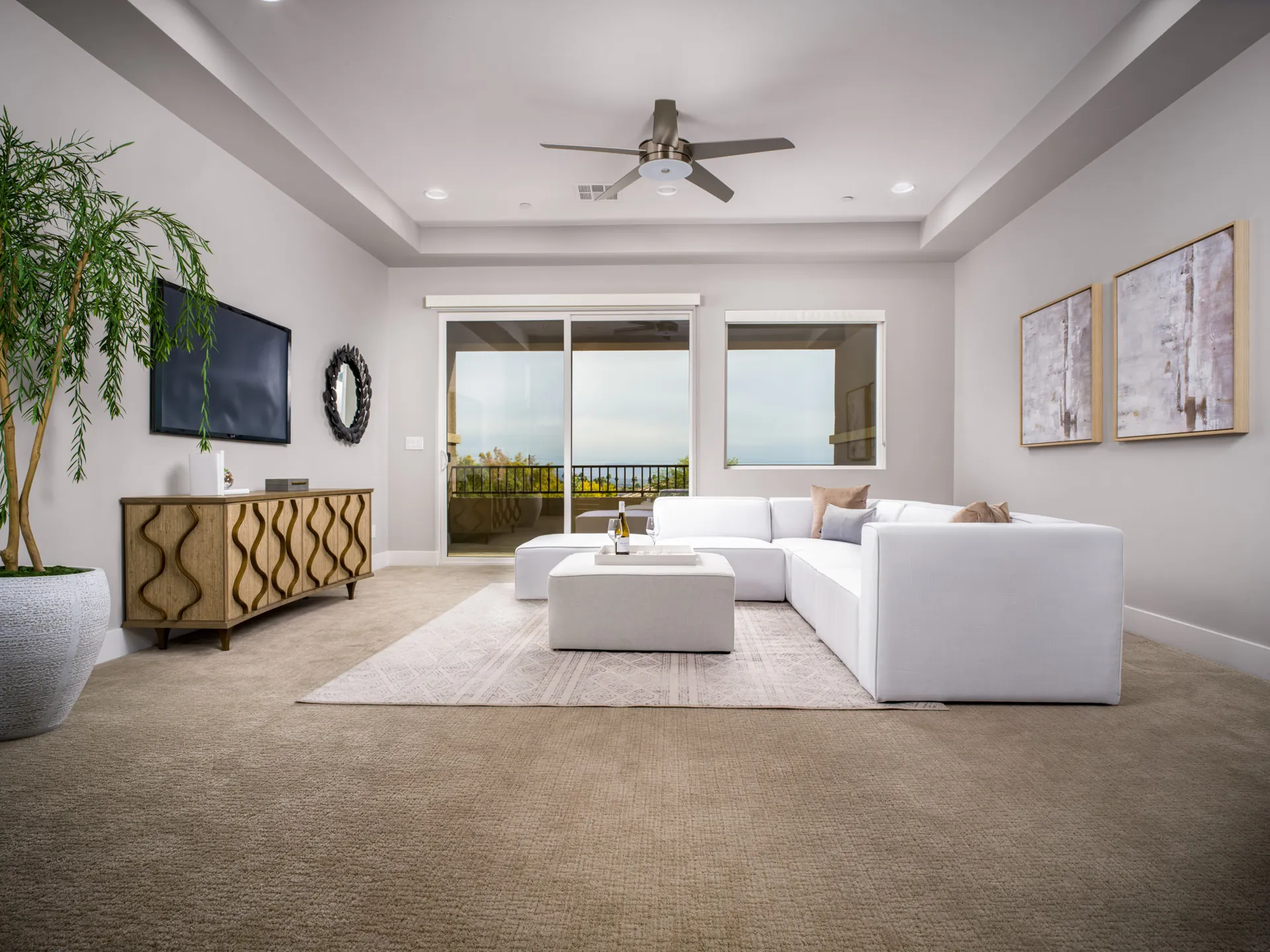 A light-filled living room with a white sectional sofa, a decorative wooden console under a wall-mounted TV, and a large potted plant near sliding glass doors.