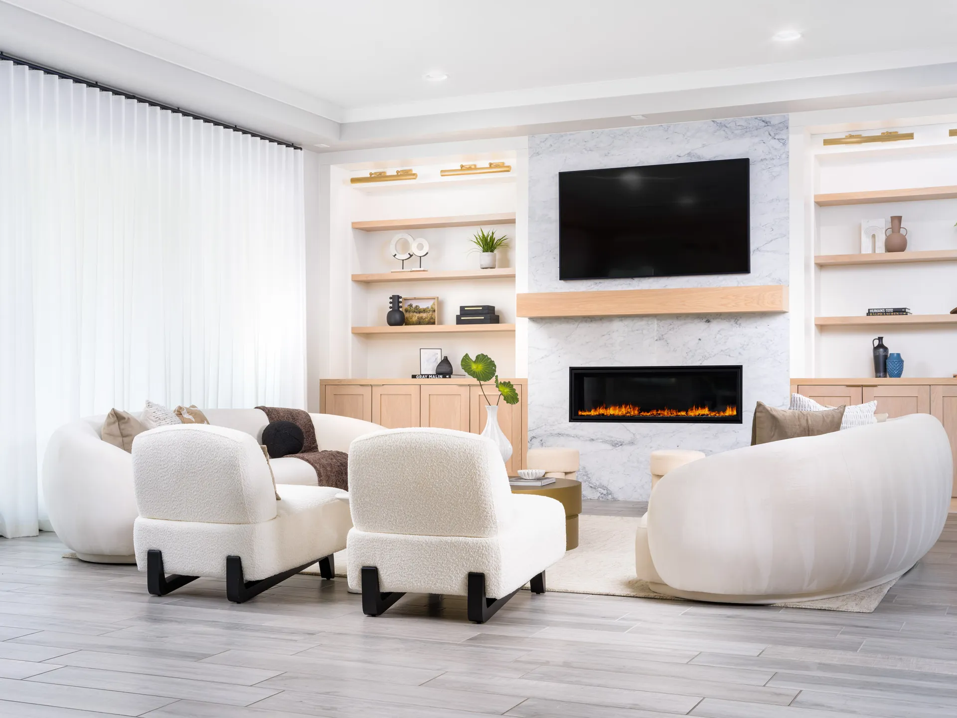 A contemporary living room featuring a marble fireplace framed by built-in shelves, curved white seating, and plush chairs in front of sheer drapes.
