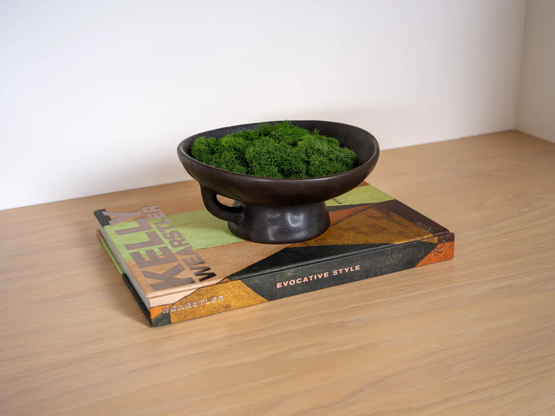 A black footed bowl filled with green moss, placed on top of a design book on a wooden shelf.