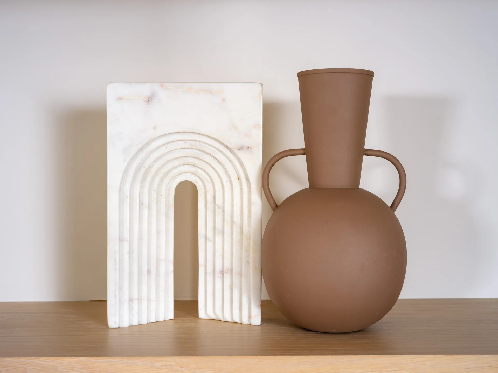 A white marble arch-shaped sculpture beside a terracotta vase on a wooden shelf.