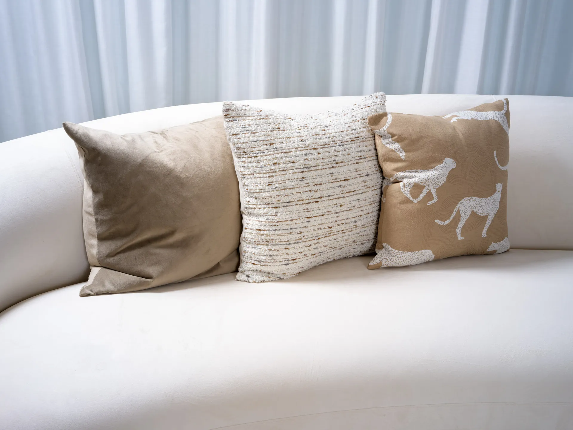 Three neutral throw pillows on a curved white sofa. One pillow features a white cheetah pattern on a tan background.