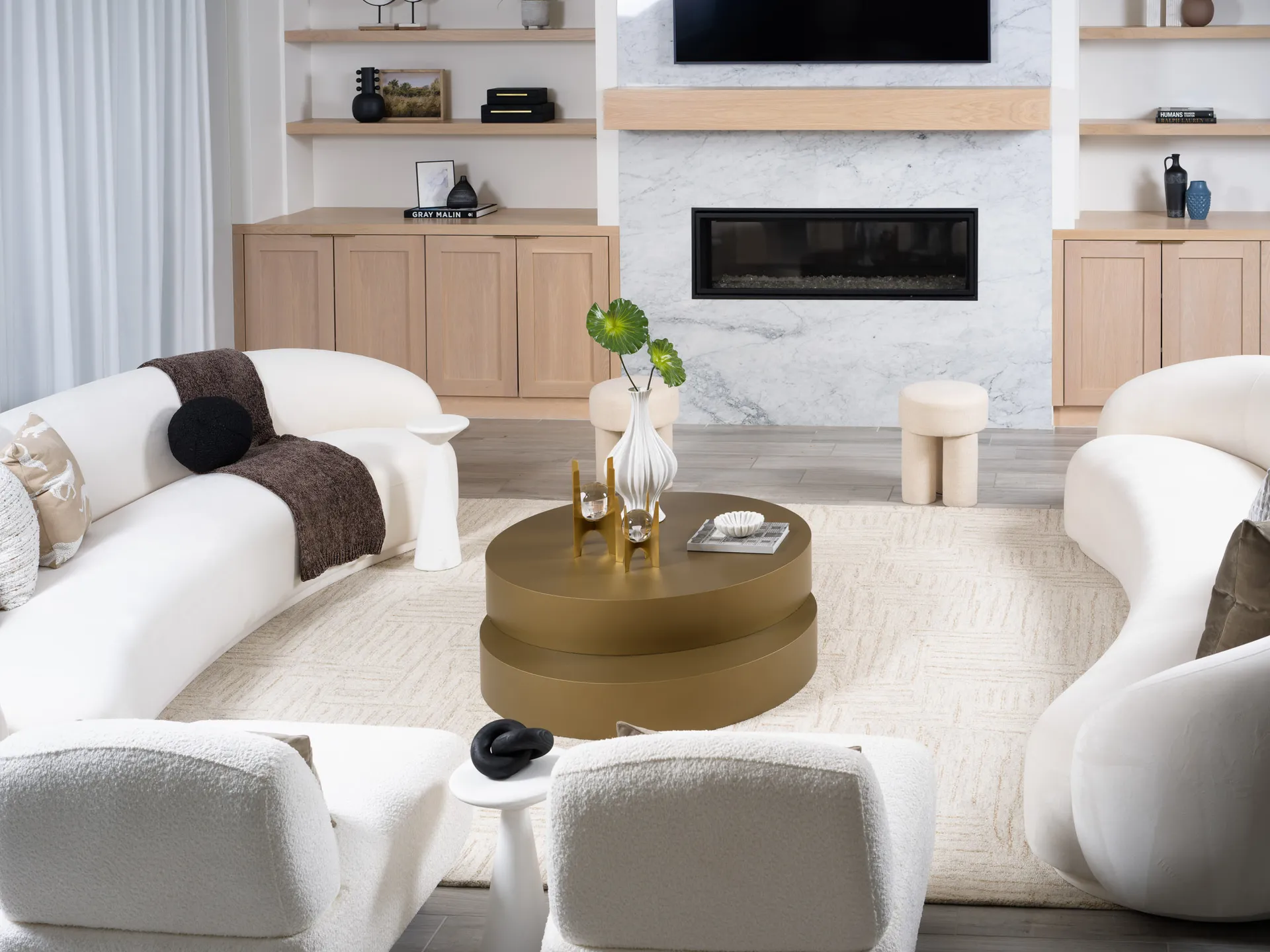 A living room featuring two curved white sofas, a round gold coffee table, and a marble fireplace with built-in cabinetry.