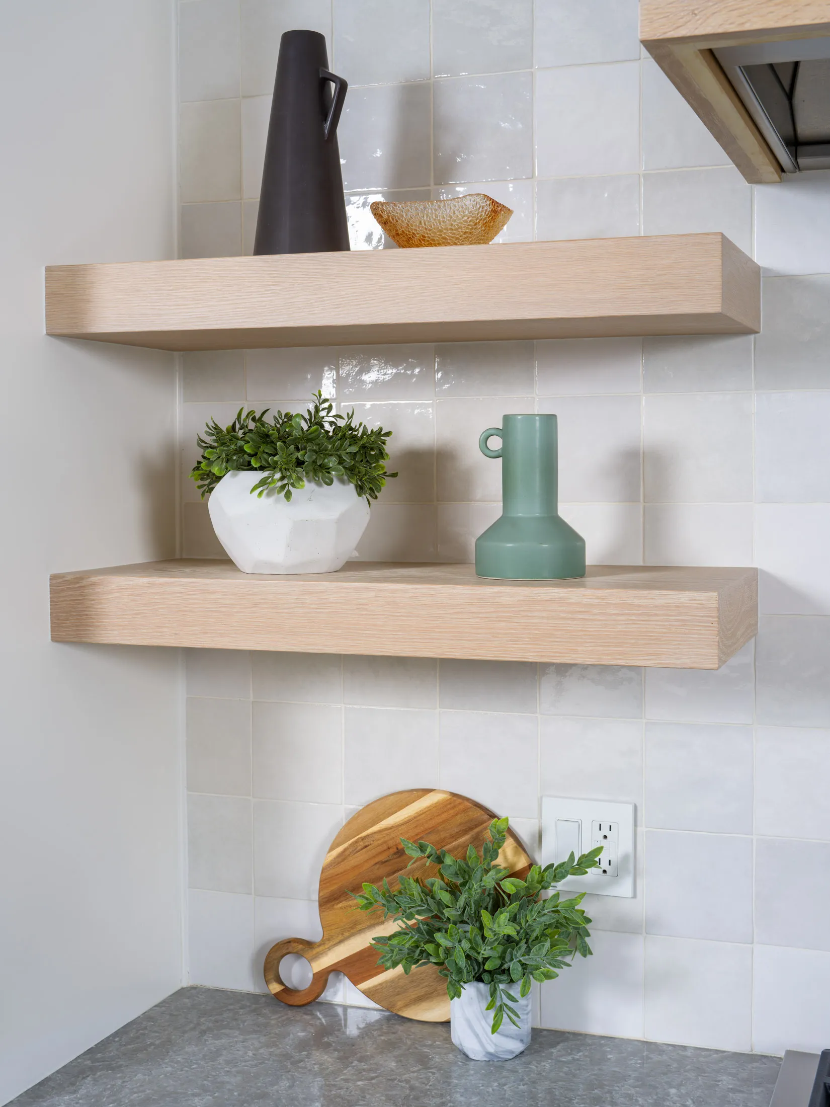 Stylish open kitchen shelves showcasing a black ceramic vase, amber decorative bowl, green geometric pitcher, and white planter with greenery, complemented by a wooden cutting board and marble plant holder on the countertop.