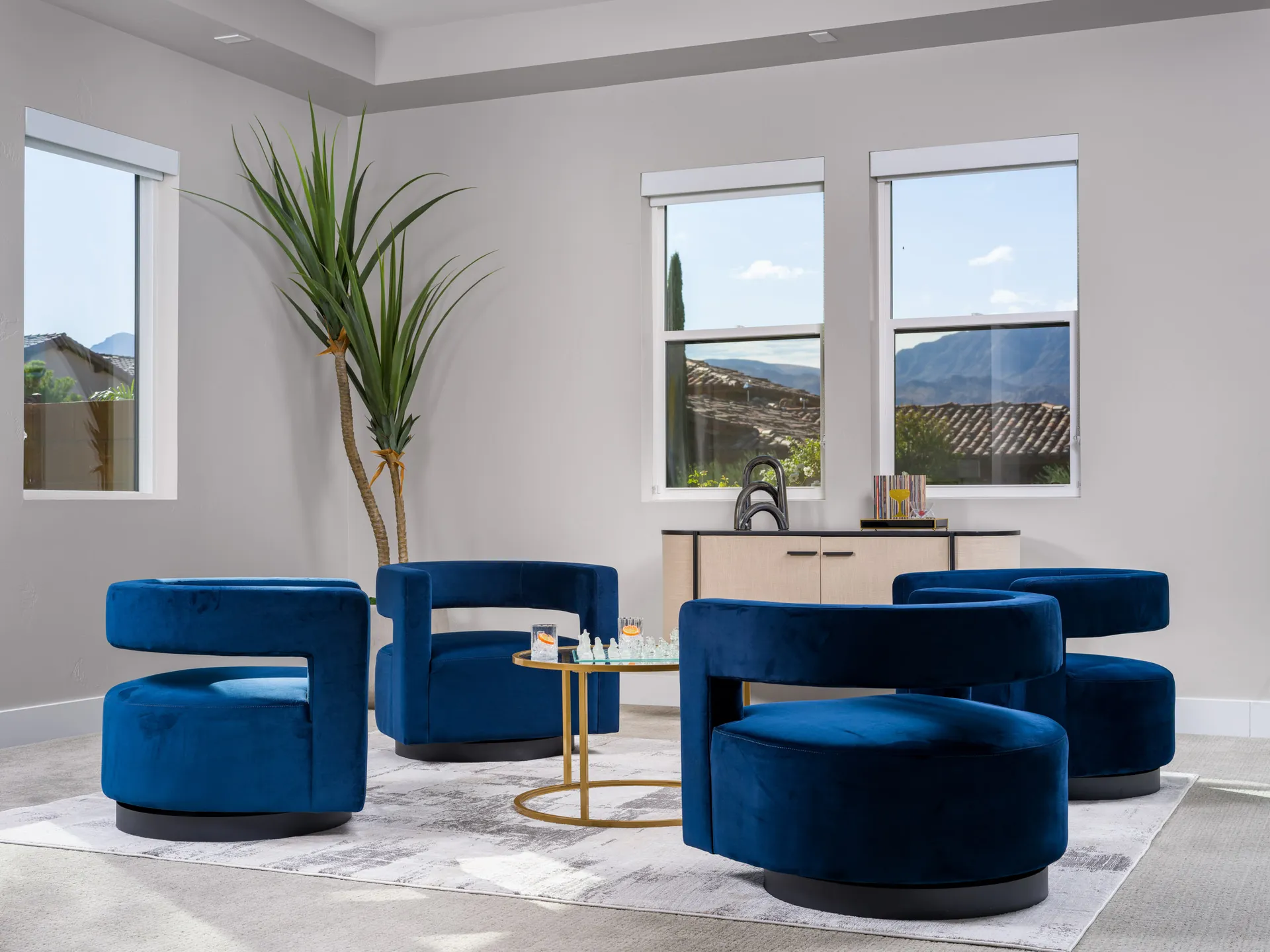 Luxury interior design featuring blue velvet swivel chairs, a sleek gold-accented coffee table, and natural light streaming through large windows, showcasing mountain views and modern decor.