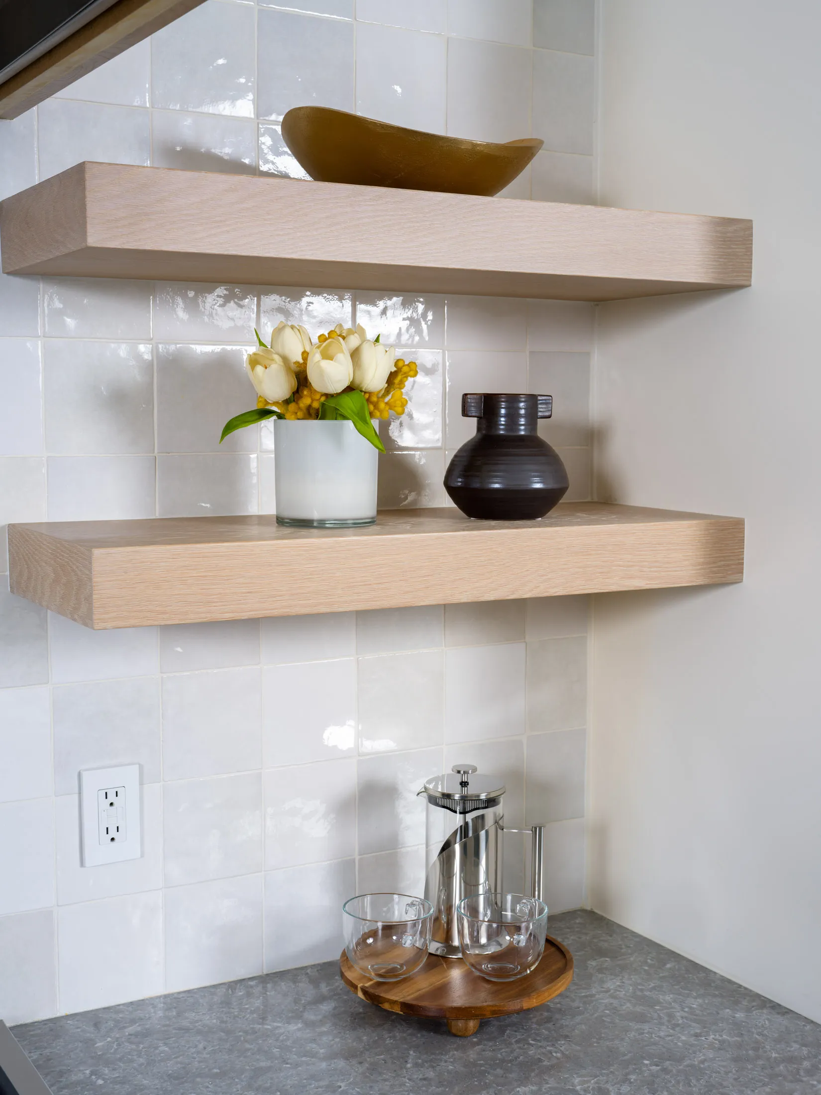Kitchen corner with open wooden shelves featuring decor items like a gold bowl, a floral arrangement in a white vase, a black ceramic vase, and a coffee set with glass cups and a French press on the counter.