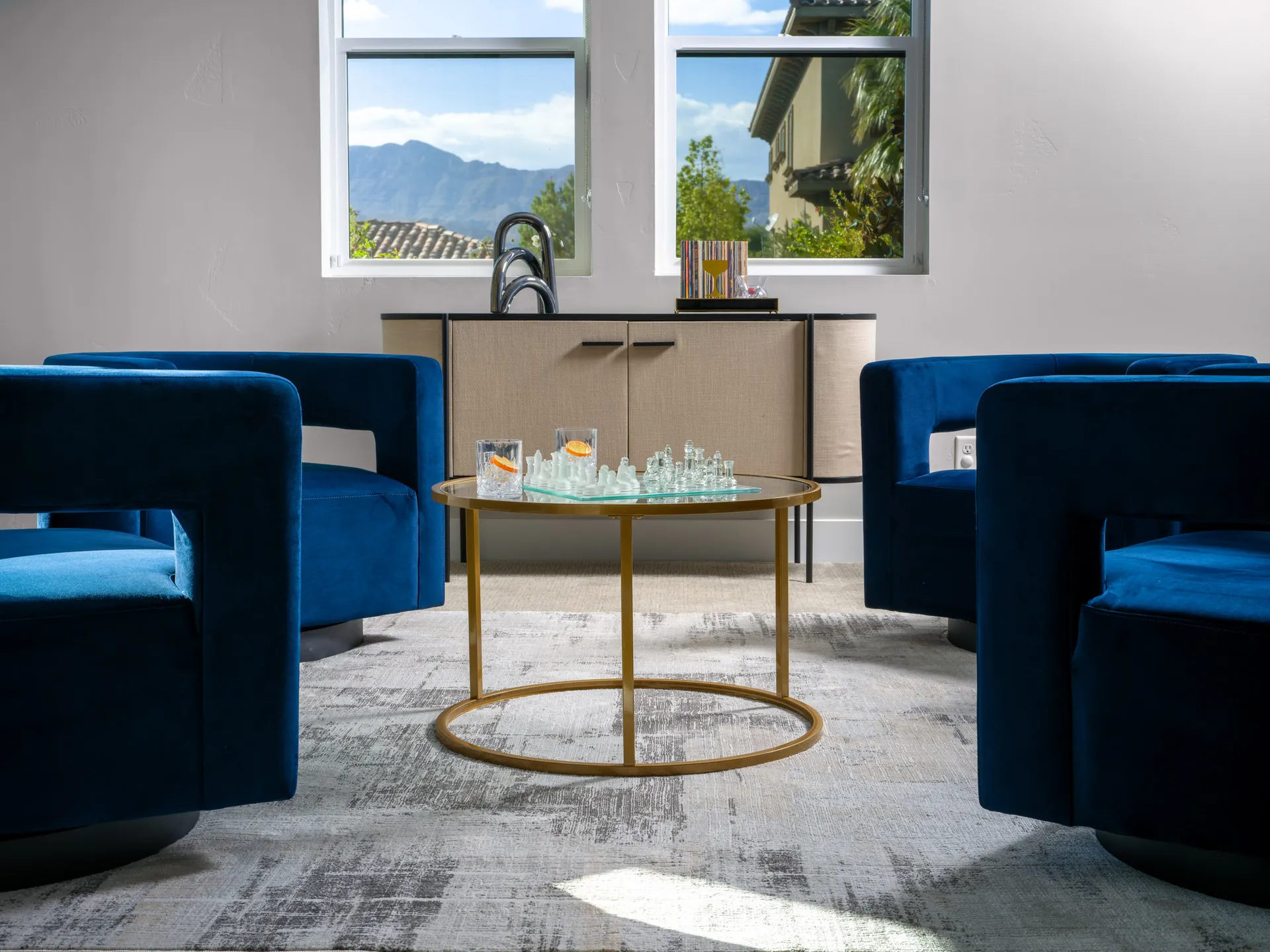 Modern sitting area featuring blue velvet armchairs, a round glass coffee table with a chess set, and a light-filled window with scenic mountain views.