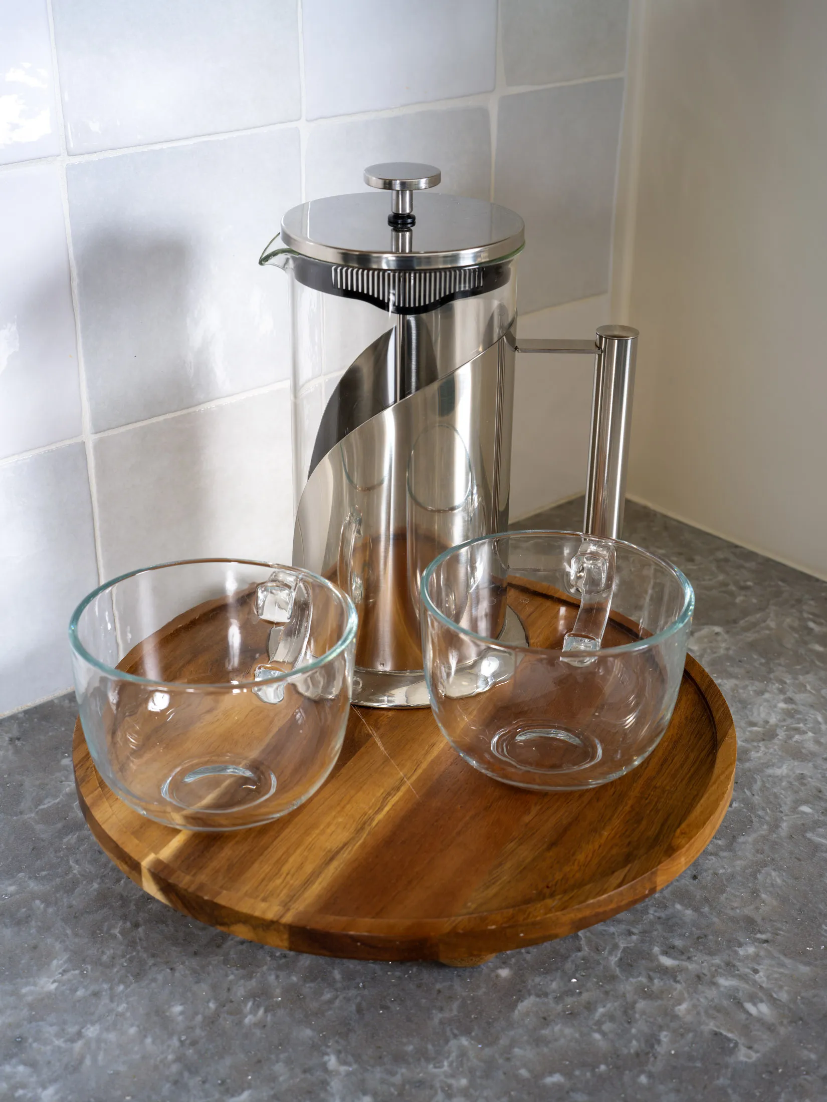 Elegant stainless steel and glass French press paired with two clear glass cups, displayed on a round wooden tray on a gray countertop.