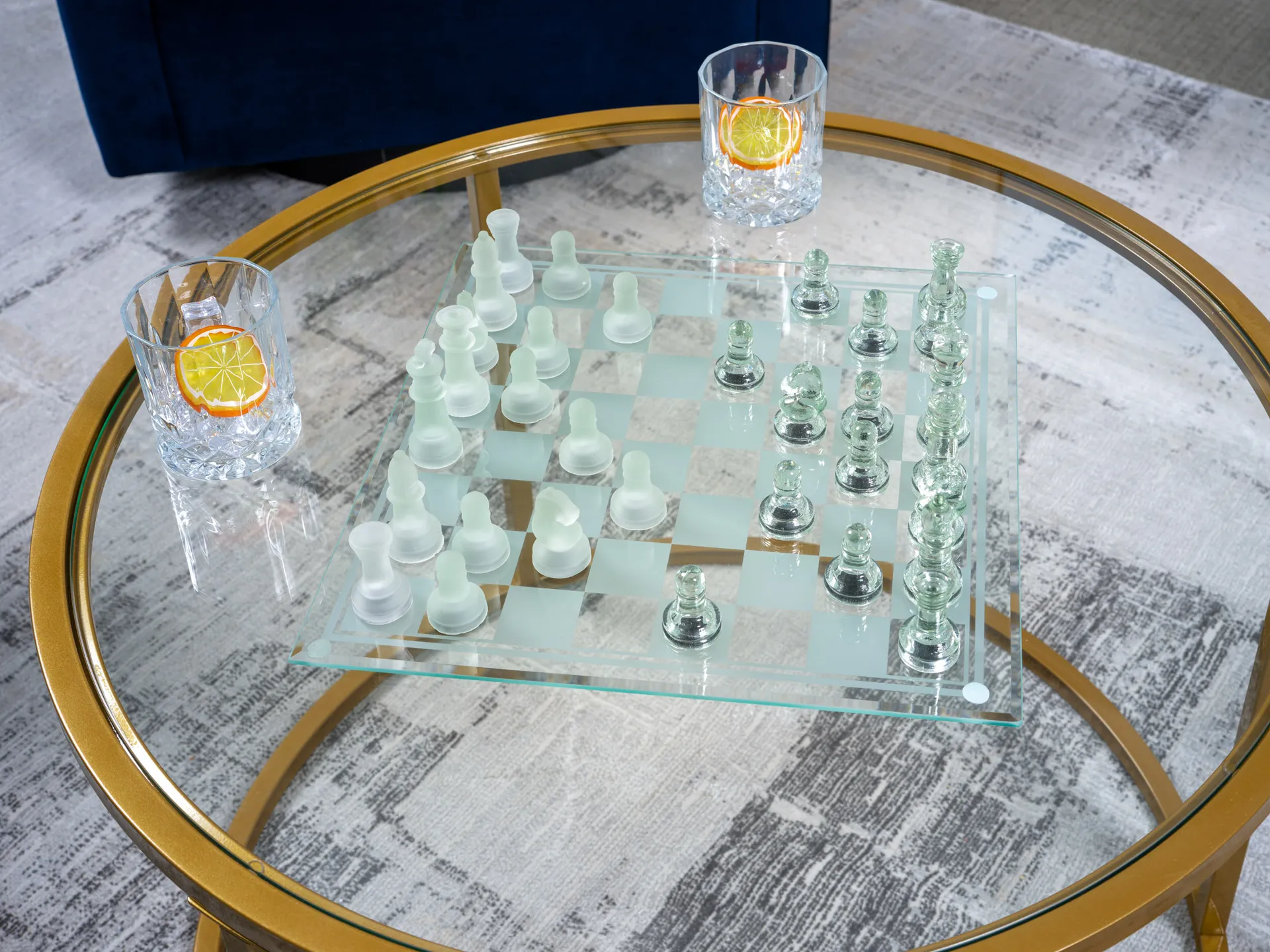 Close-up of a glass chess set on a round gold-framed table, flanked by two crystal glasses with citrus garnishes.