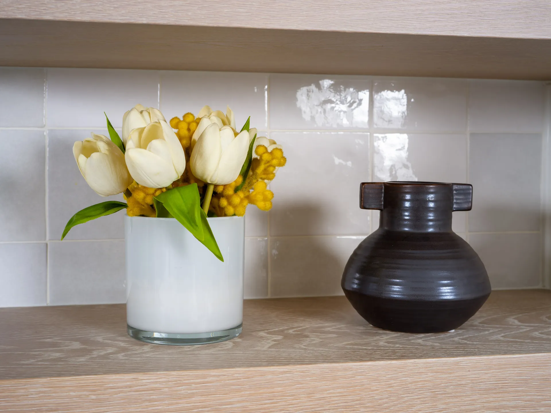 Decorative shelf featuring a white vase with yellow and white tulips alongside a black ceramic vase with unique handles.