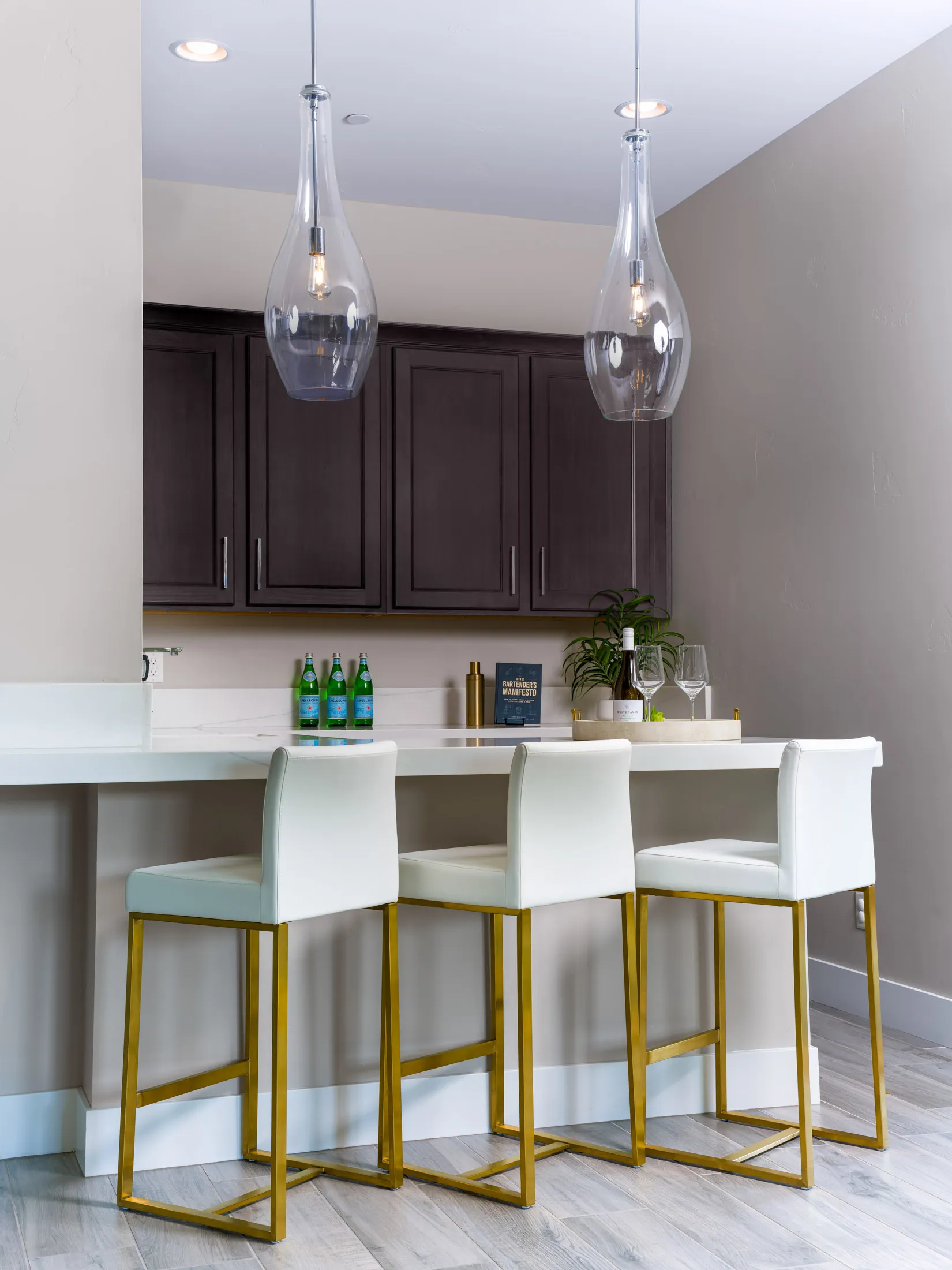Contemporary kitchen island featuring three white barstools with gold legs, illuminated by glass pendant lights, against dark wood cabinetry.
