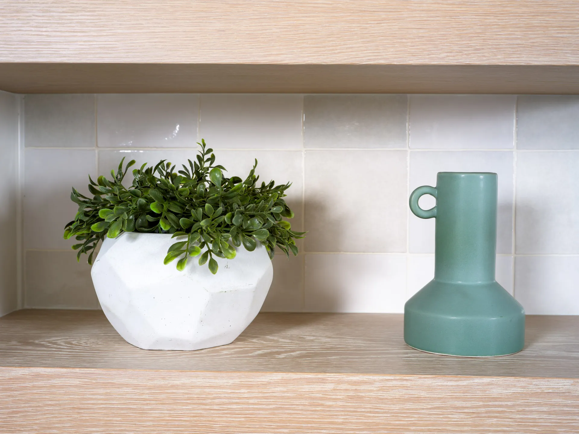 A white geometric planter with lush greenery next to a matte green ceramic vase, styled on a light wood shelf with a tiled backsplash.