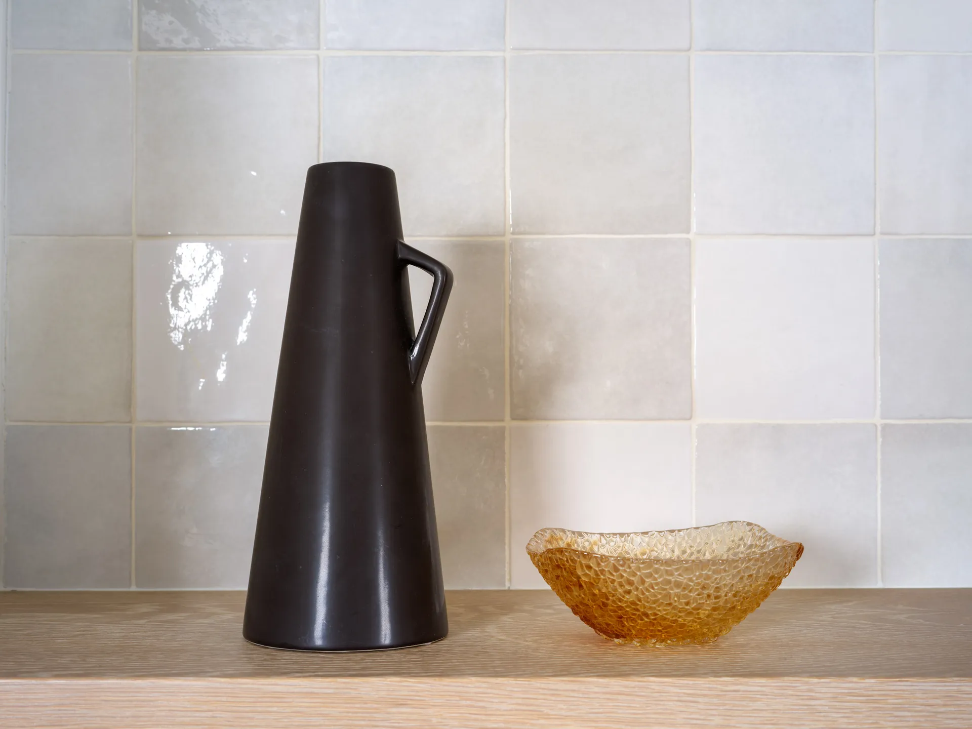 A matte black vase with a handle paired with a textured amber glass bowl, set against a light tiled backsplash on a wooden shelf.