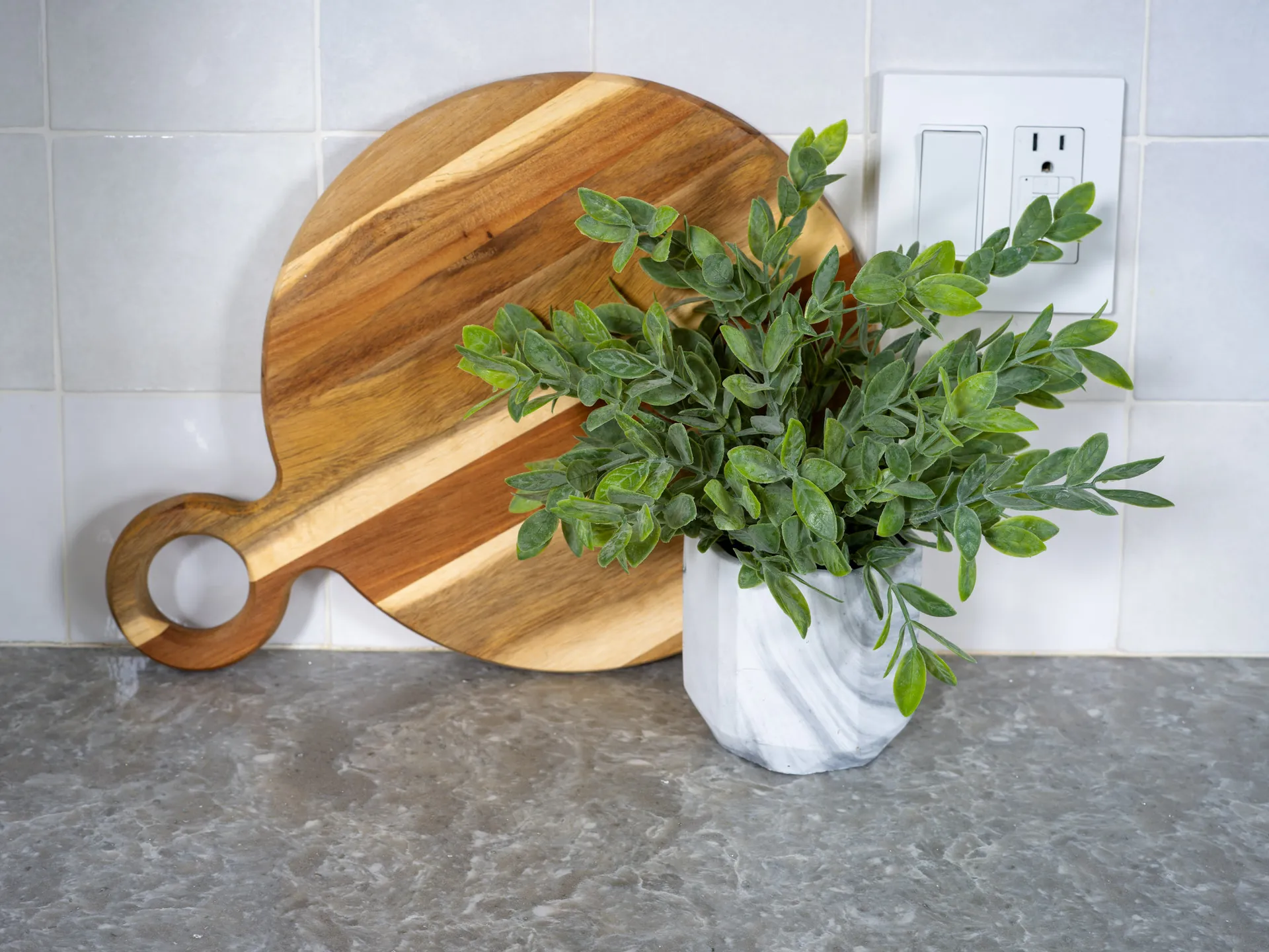 A natural wood cutting board with a handle, paired with a lush green plant in a marble-textured vase, set on a gray countertop with tiled backsplash.