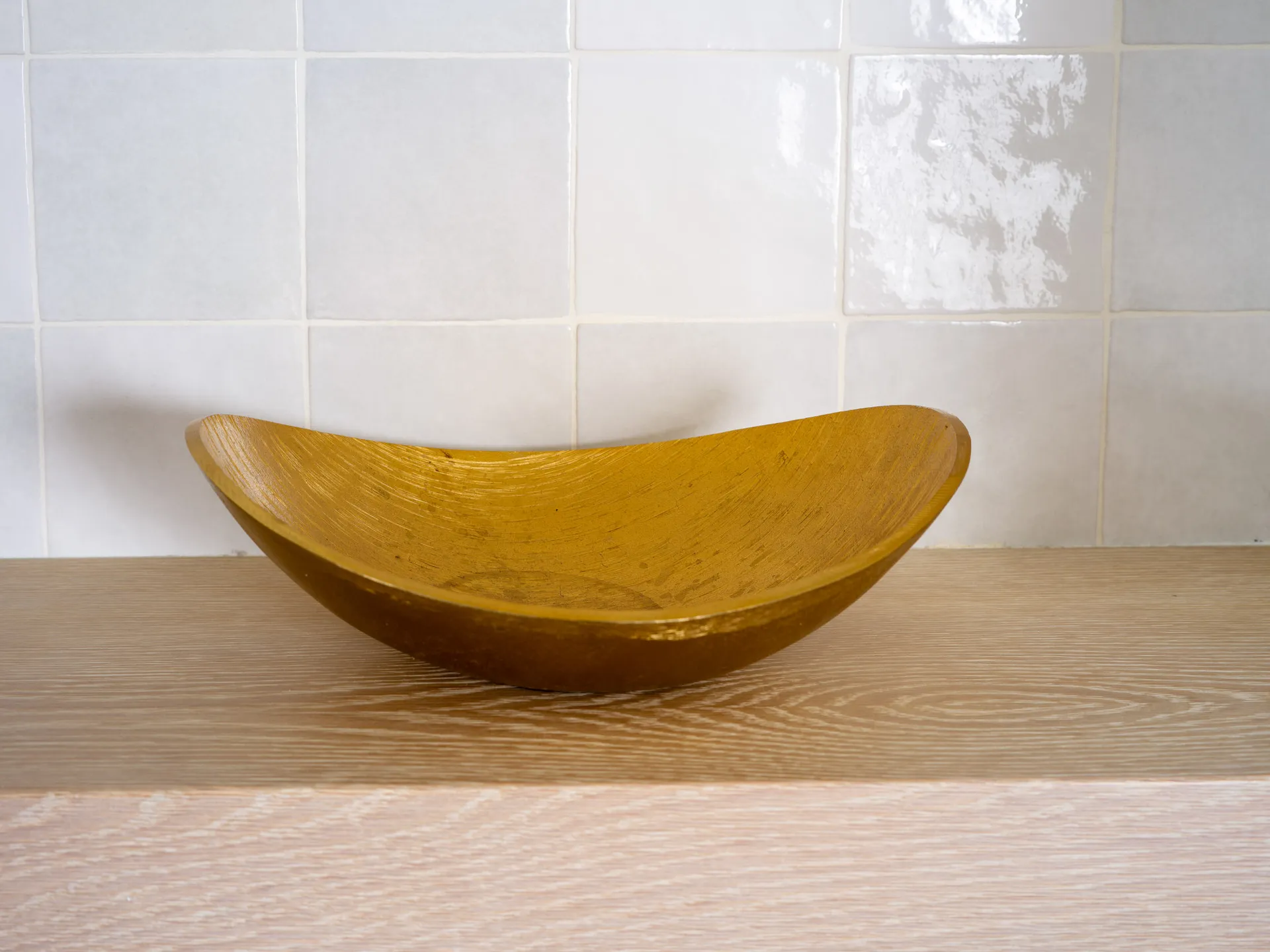 A textured golden bowl with a sleek design placed on a light wooden surface against a tiled backdrop.