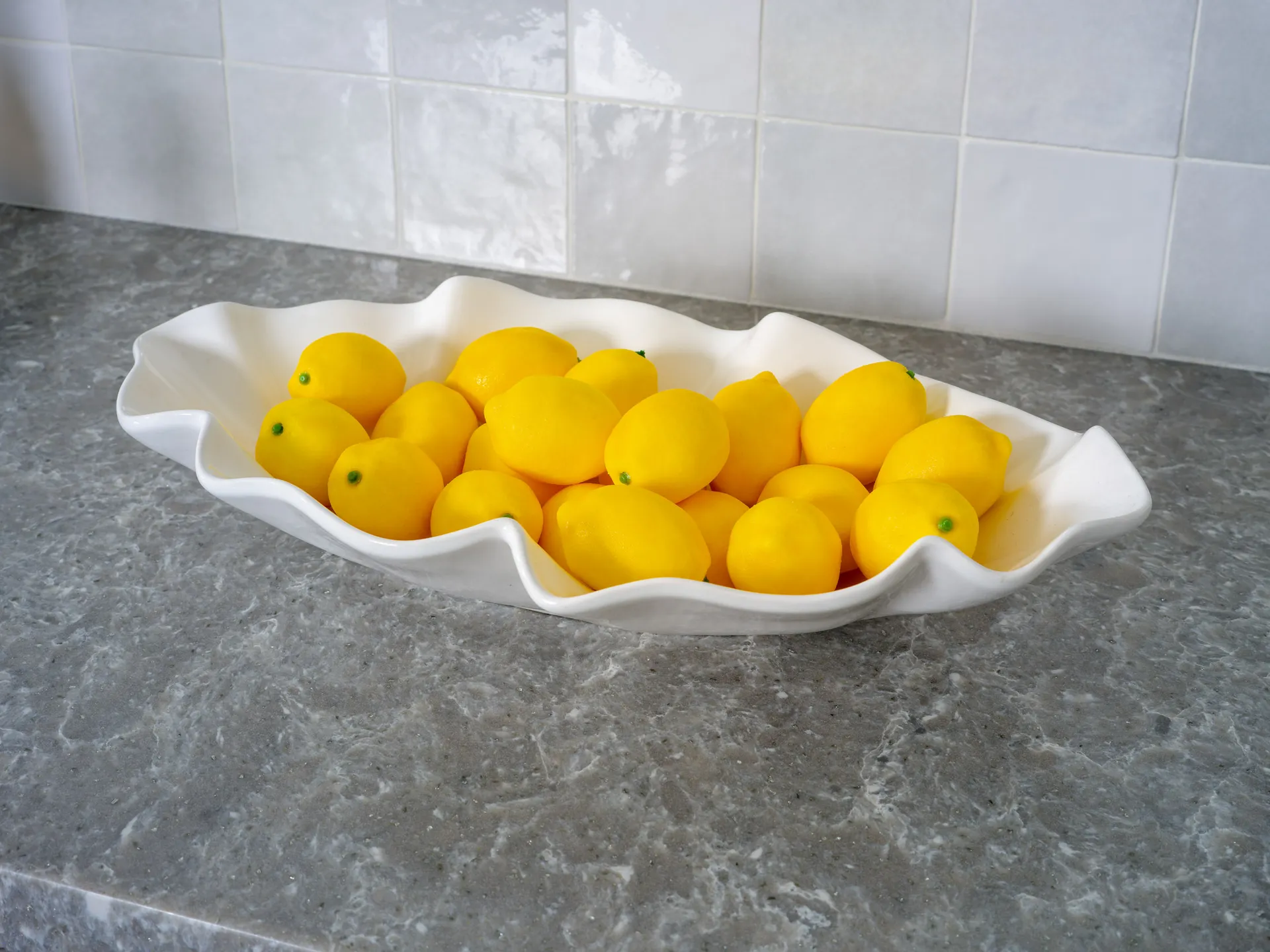 A sculpted white bowl filled with bright yellow lemons, set on a gray marble countertop with a tiled backsplash.