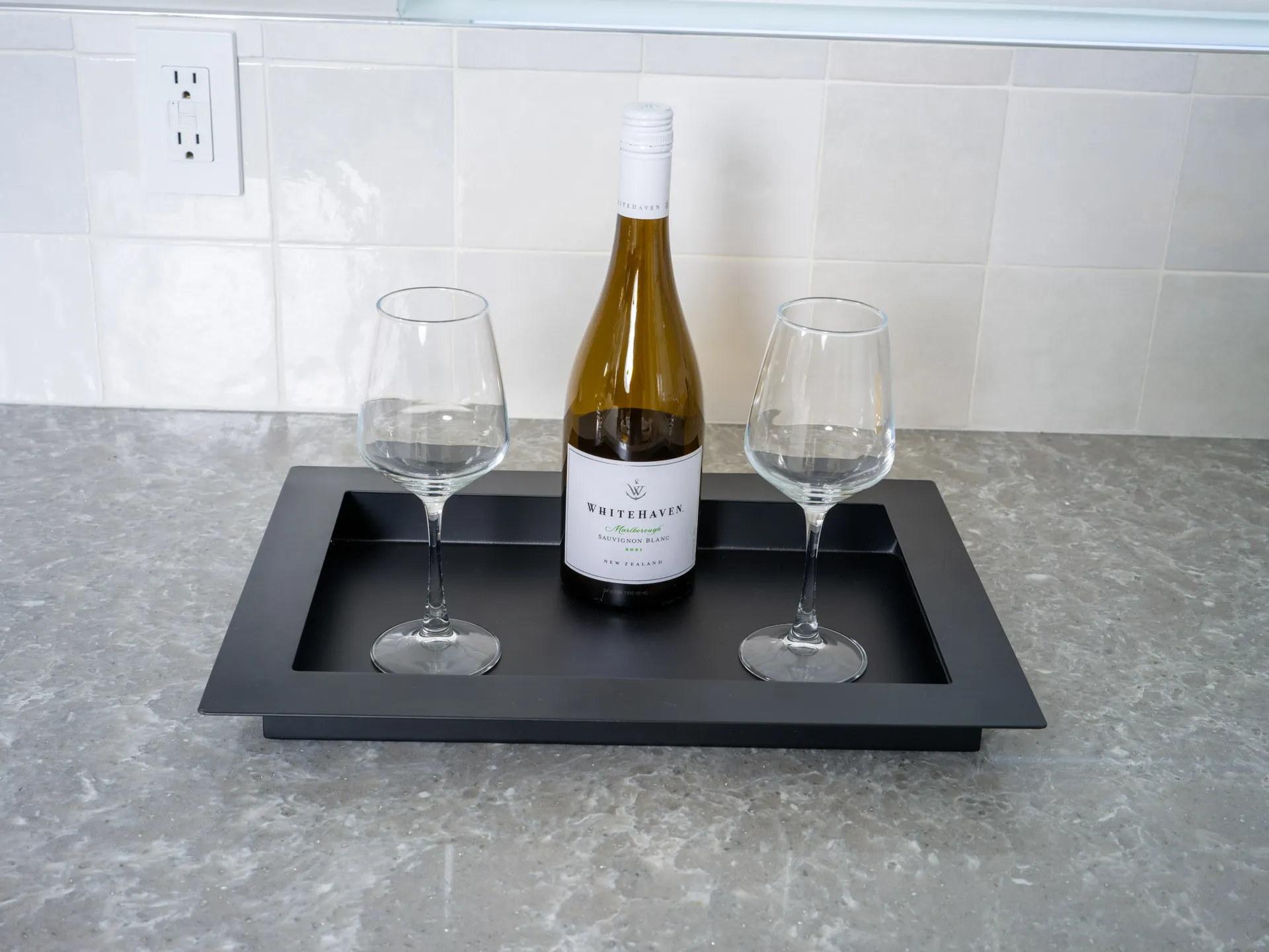 A minimalist black tray holding a bottle of Whitehaven Sauvignon Blanc and two empty wine glasses, placed on a gray marble countertop against a tiled backsplash.