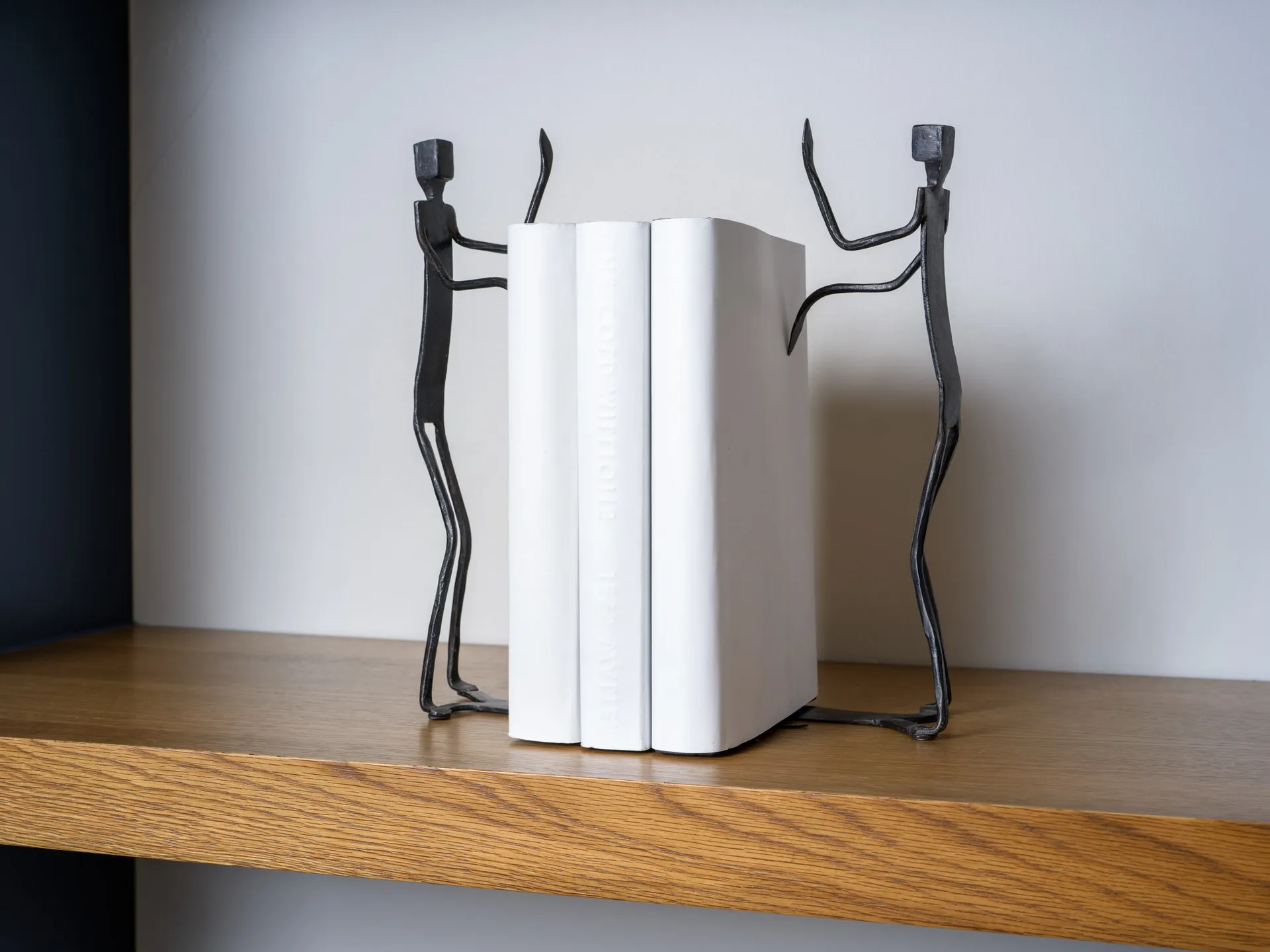 Two abstract iron sculptures resembling human figures holding white books on a wooden shelf.