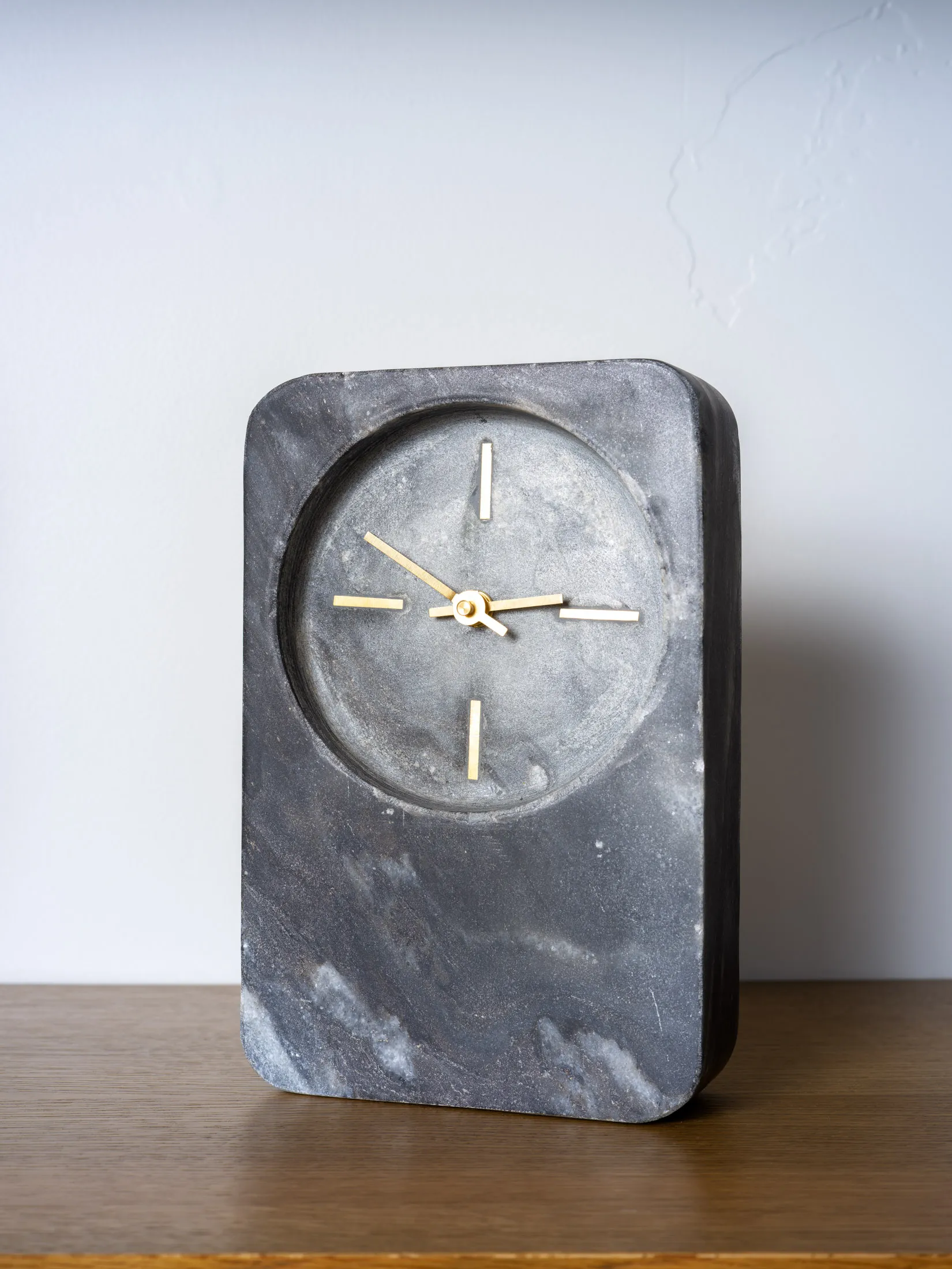 A sleek rectangular marble clock with subtle veining and minimalist gold hands, placed on a wooden shelf.