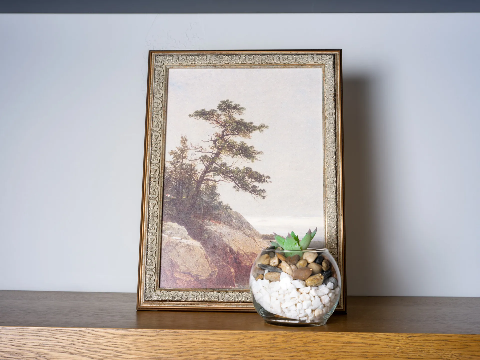 A framed landscape painting of a serene tree by a rocky shore, paired with a glass terrarium holding white pebbles, stones, and a green succulent.