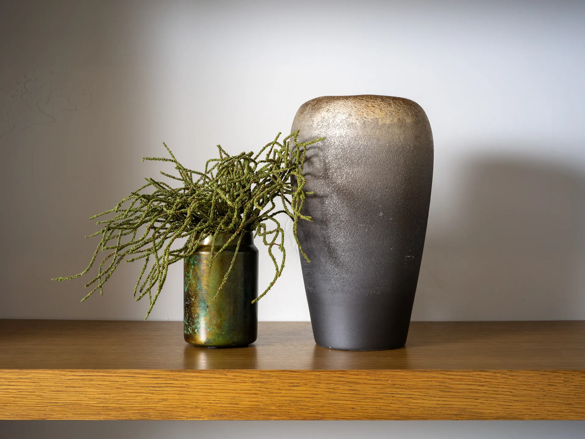 A textured black gradient vase paired with a smaller iridescent vase holding greenery, styled on a wooden shelf.