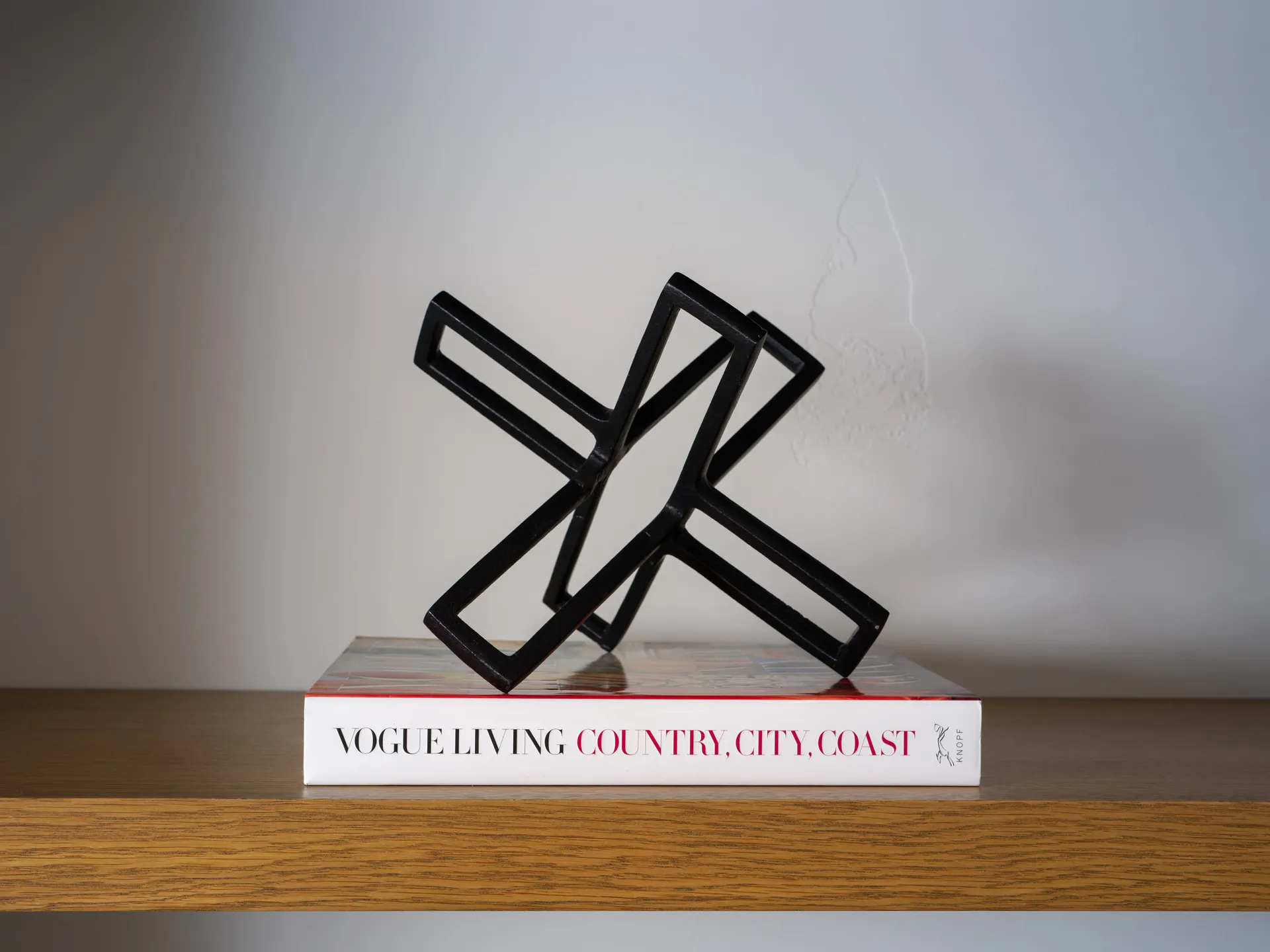 A sleek black geometric sculpture resting atop the book ‘Vogue Living: Country, City, Coast’ on a wooden shelf