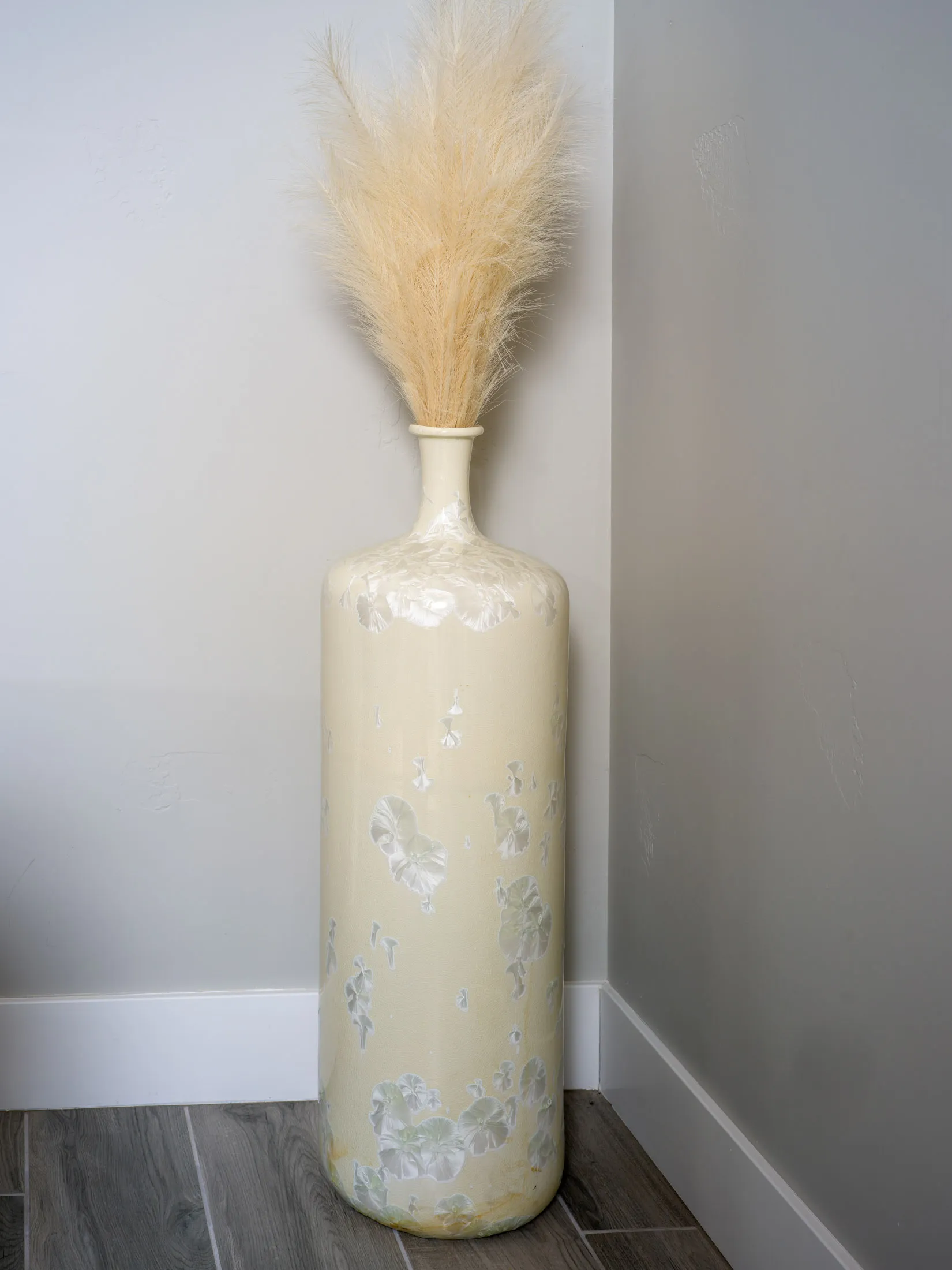 A tall beige ceramic vase with delicate floral patterns, filled with fluffy pampas grass, placed in the corner of a modern interior with wood flooring.