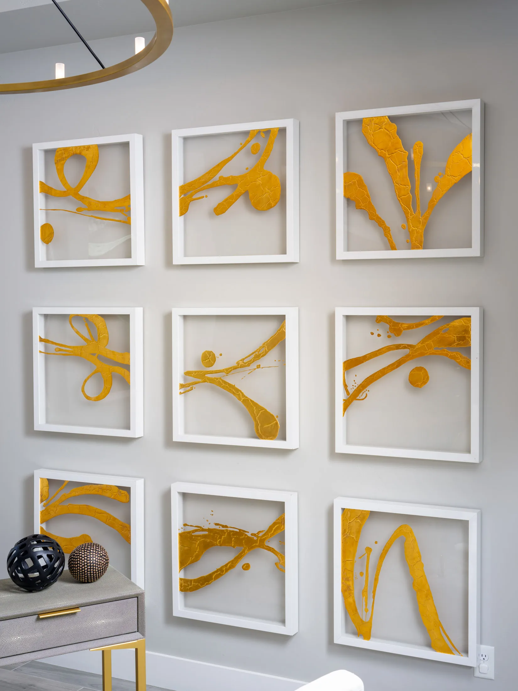 Nine framed pieces of abstract gold artwork arranged in a grid pattern on a light gray wall, with a modern console table below featuring decorative spheres.
