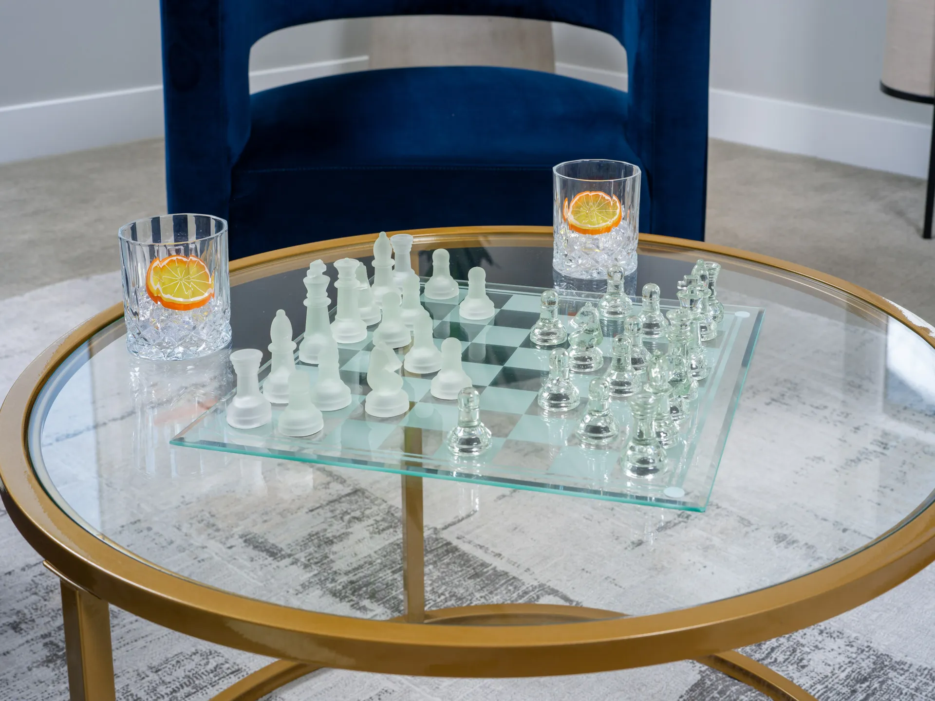 A frosted glass chess set on a round gold coffee table, accompanied by two crystal glasses garnished with orange slices, placed on a neutral textured rug