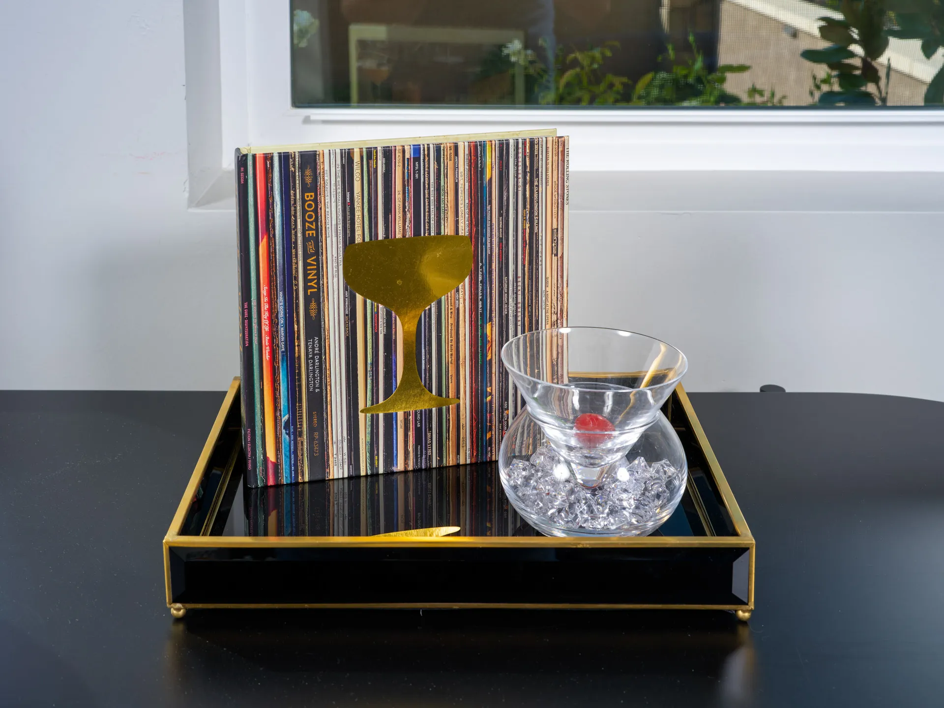 A chic gold-trimmed tray showcasing a clear cocktail glass with ice and a cherry, alongside a vibrant vinyl-themed decor piece, placed on a sleek black surface.