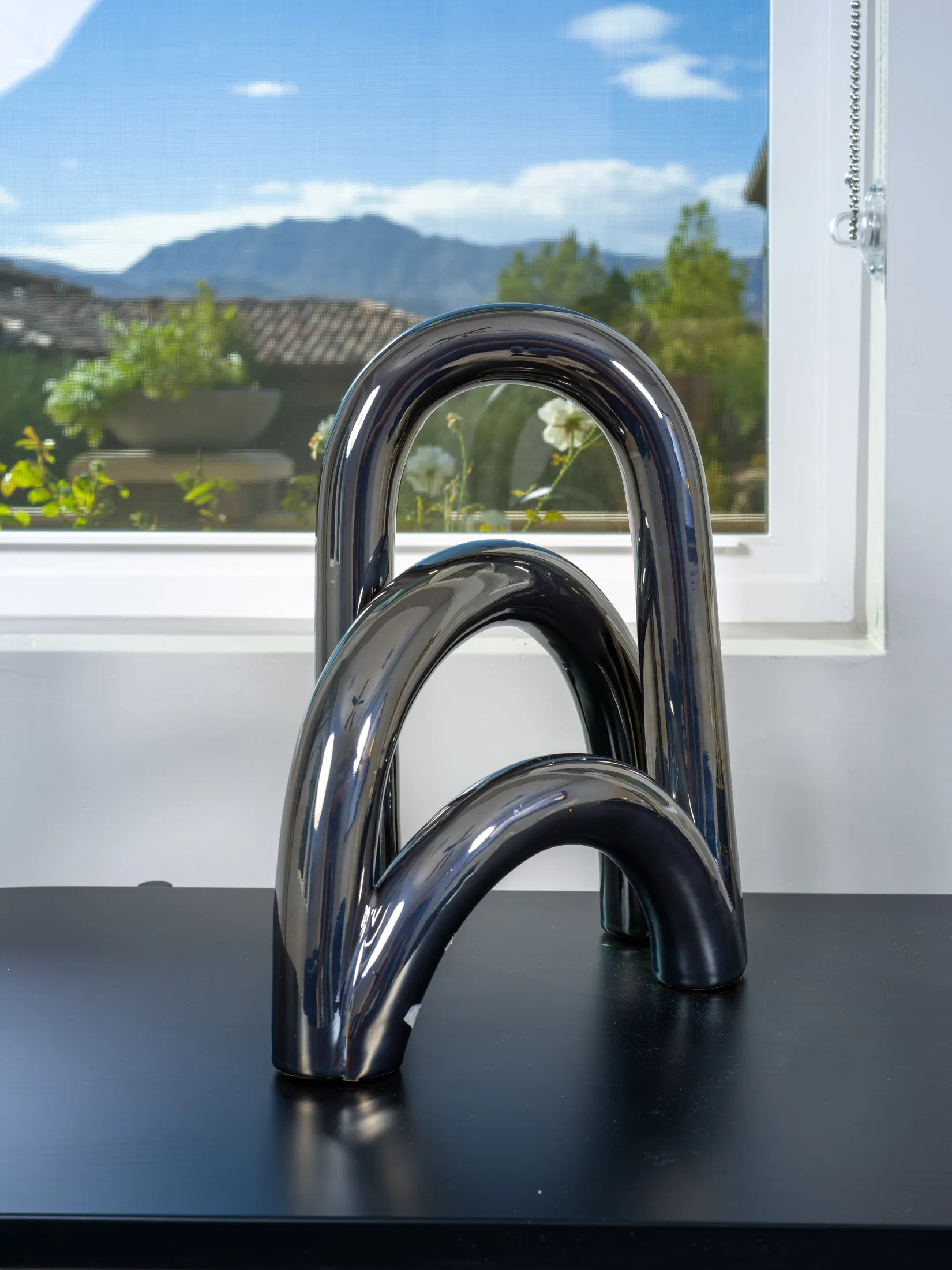 A sleek chrome accent sculpture displayed on a black table, with a scenic mountain view framed by a window in the background.