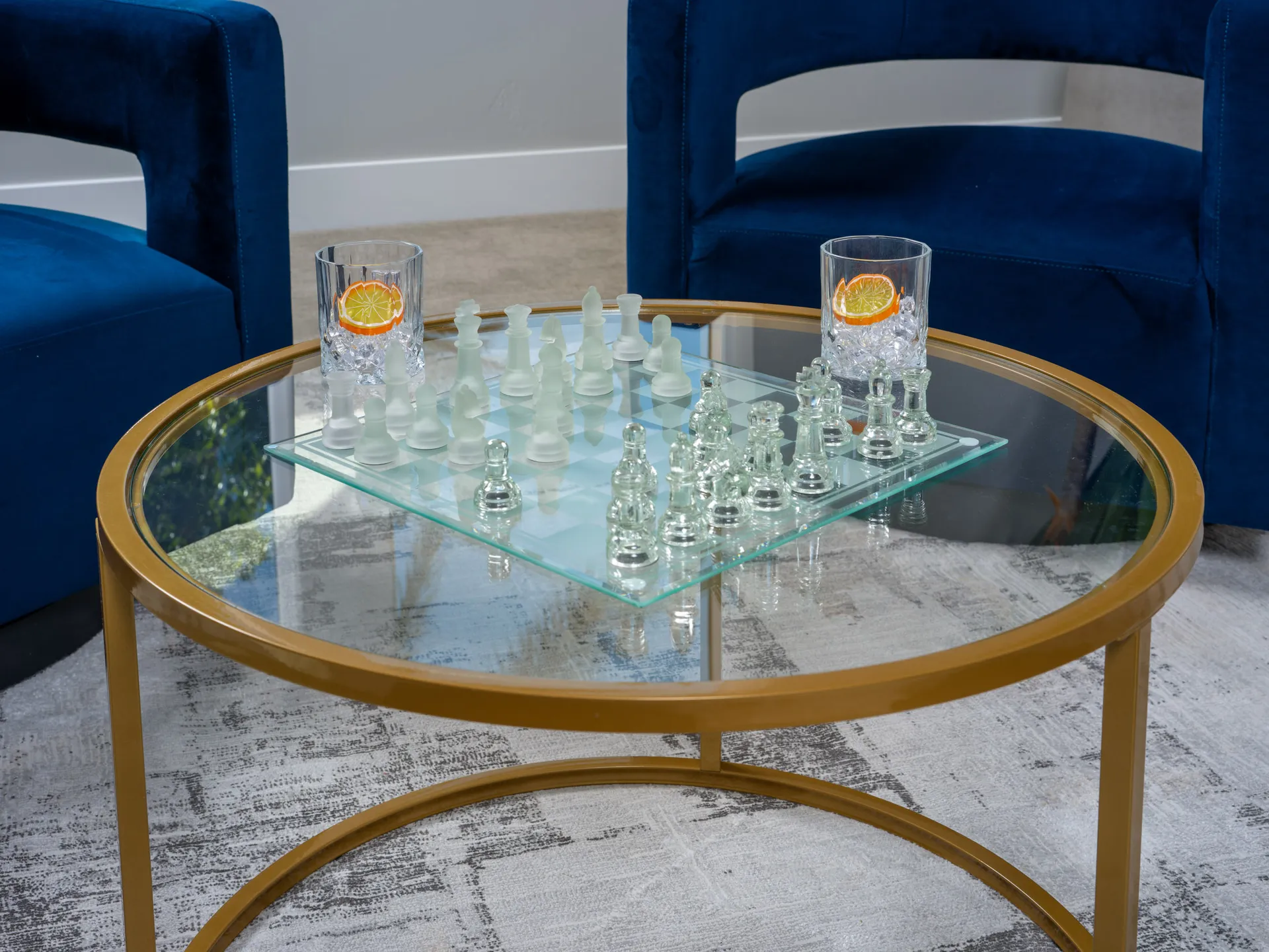 A frosted glass chess set displayed on a round glass coffee table with gold trim, paired with luxurious blue chairs in a stylish setting.
