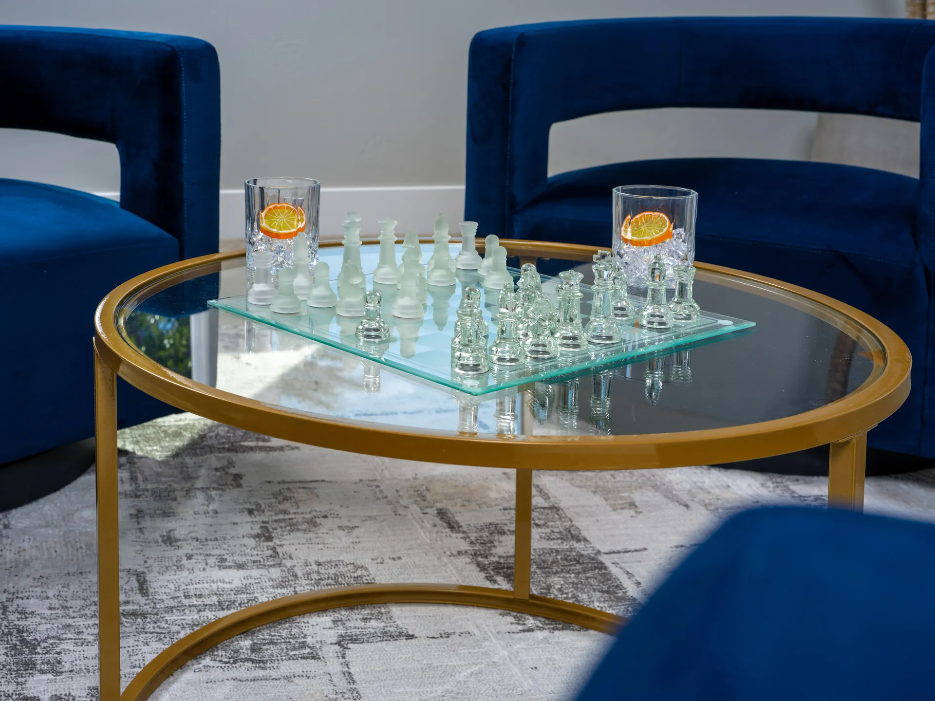 Close-up of a frosted glass chess set on a round gold coffee table, surrounded by plush blue chairs in a luxury-staged room.
