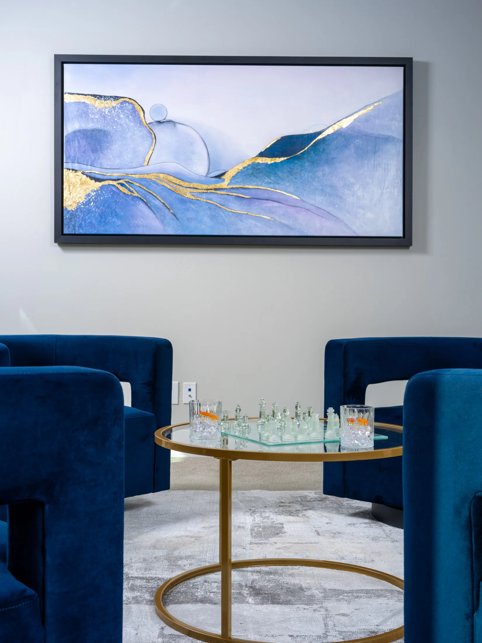 Modern lounge area featuring deep blue velvet chairs, a glass chess table with drinks, and a gold-accented abstract painting styled for luxury home staging by Stately.