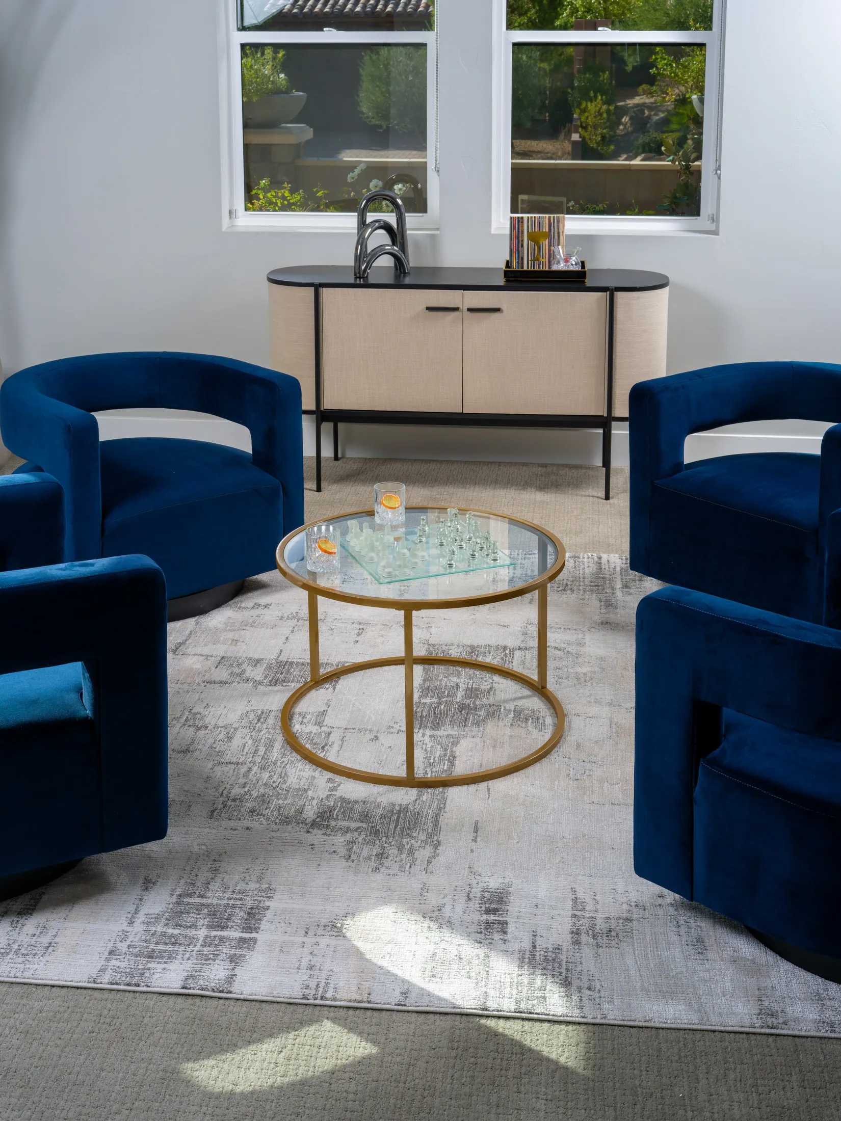 Modern sitting area featuring rich blue velvet chairs, a gold-framed glass coffee table with a chess set, and a beige console with a built-in sink, styled for luxury home staging by Stately.