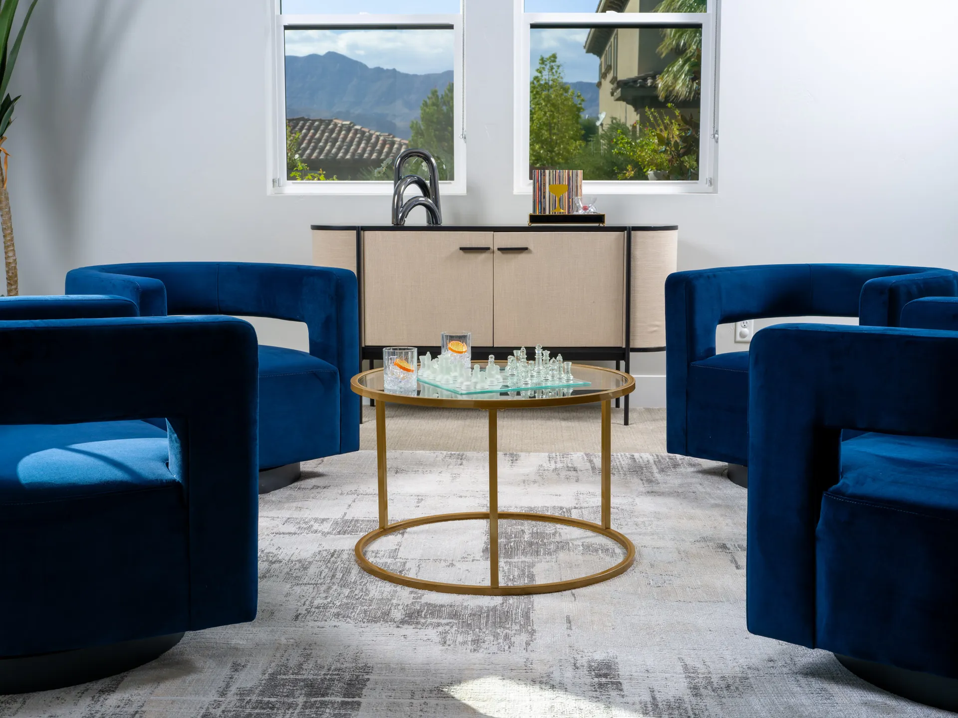 Elegant sitting area featuring bold blue velvet chairs, a gold circular coffee table with a chess set, and a minimalist beige sideboard against a backdrop of scenic mountain views. Styled by Stately for luxury home staging.