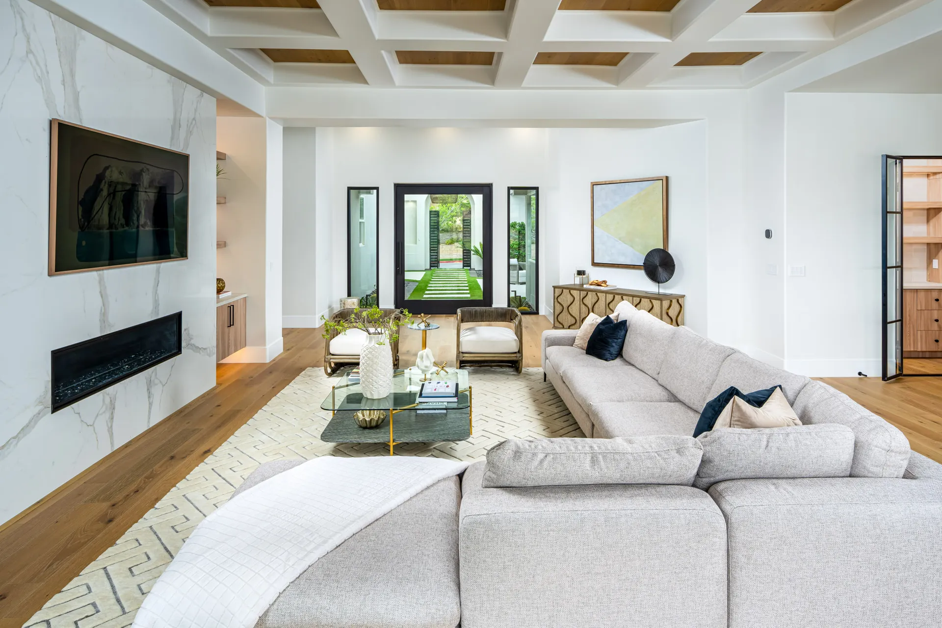 A large sectional sofa faces a marble fireplace and glass coffee table. A coffered ceiling and an outdoor walkway are visible in the background.