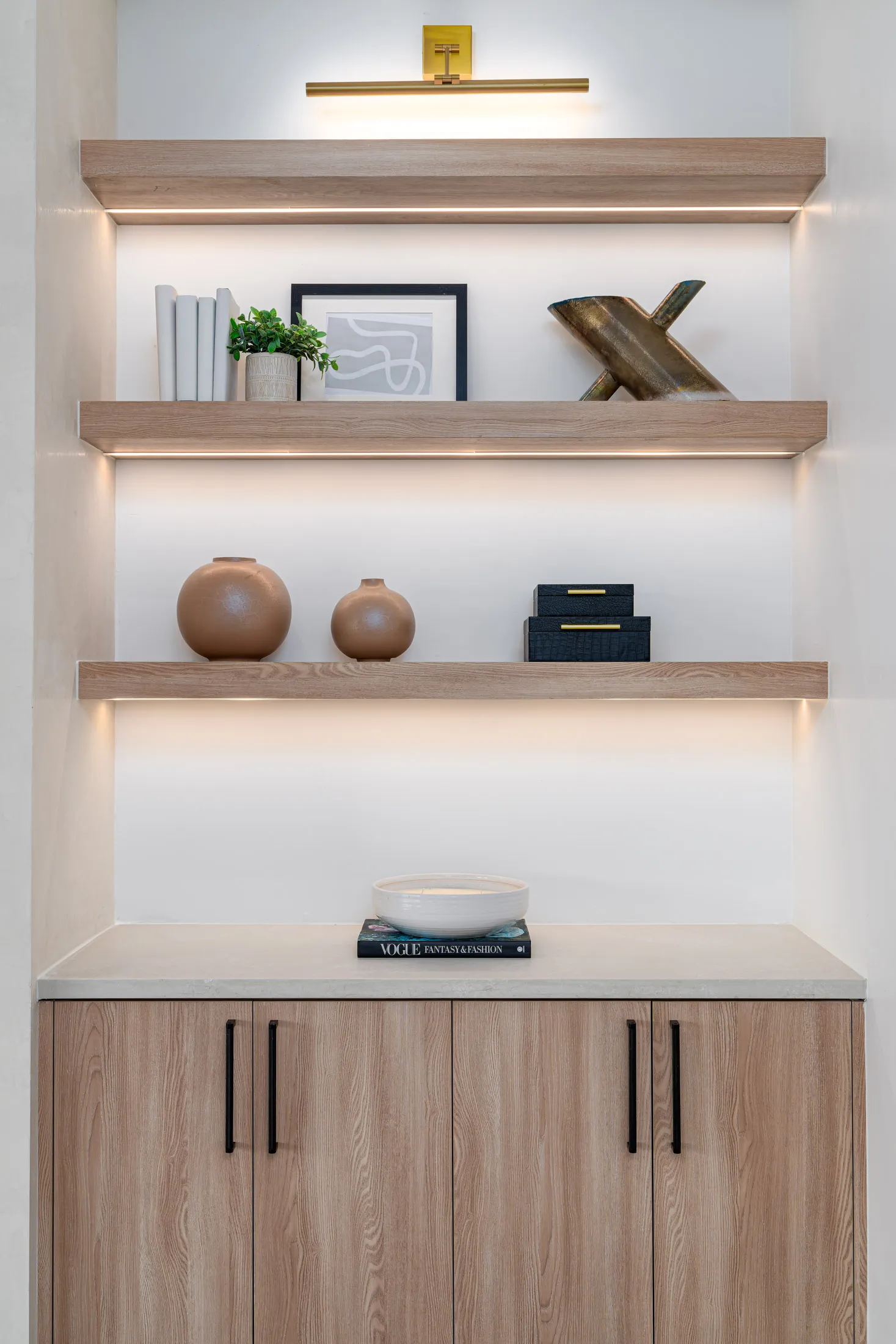 Wooden floating shelves illuminated by built-in lights, displaying round vases, boxes, and a framed artwork, under a brass picture light.